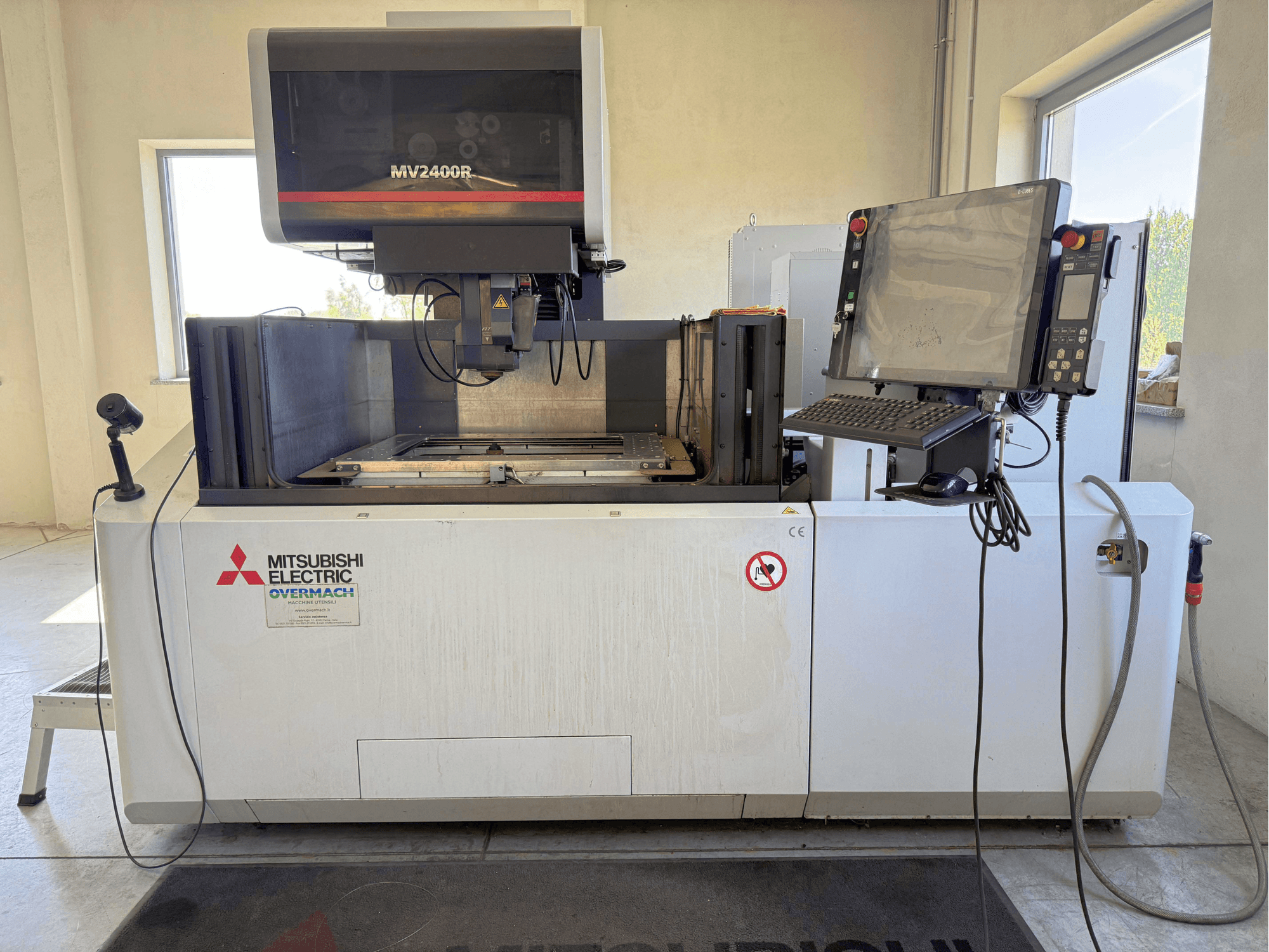 Mitsubishi MV2400R traat-EDM-masin eestvaates, millel on juhtpaneel, tööala ja ohutusmärgistus.