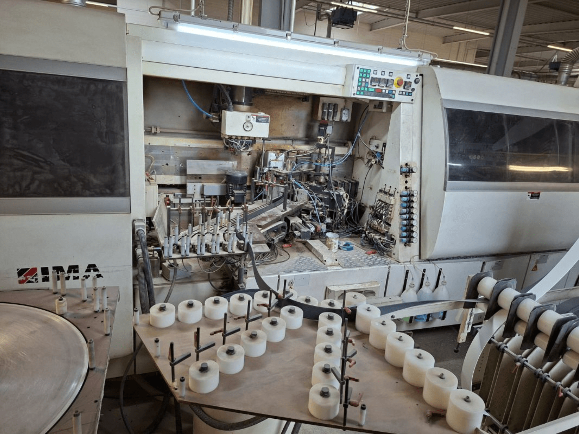 IMA masinad küljelt vaadatuna, kus näidatakse keerulisi mehaanilisi komponente ja ümmargune laud koos valgete poolidega.