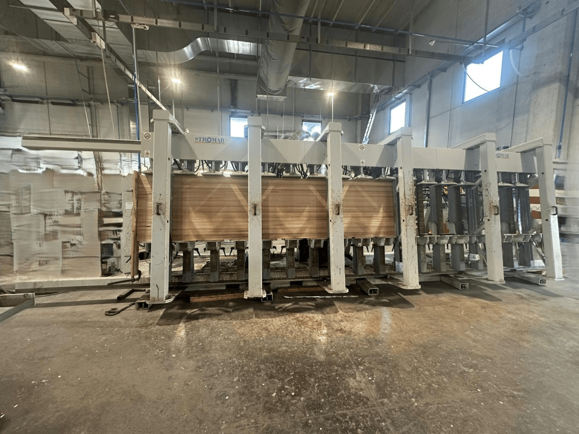 Tööstuslik STROMABi masin laos, esiplaanil, töödeldavad puitpaneelid, metallkonstruktsioon ja mehaanilised osad.