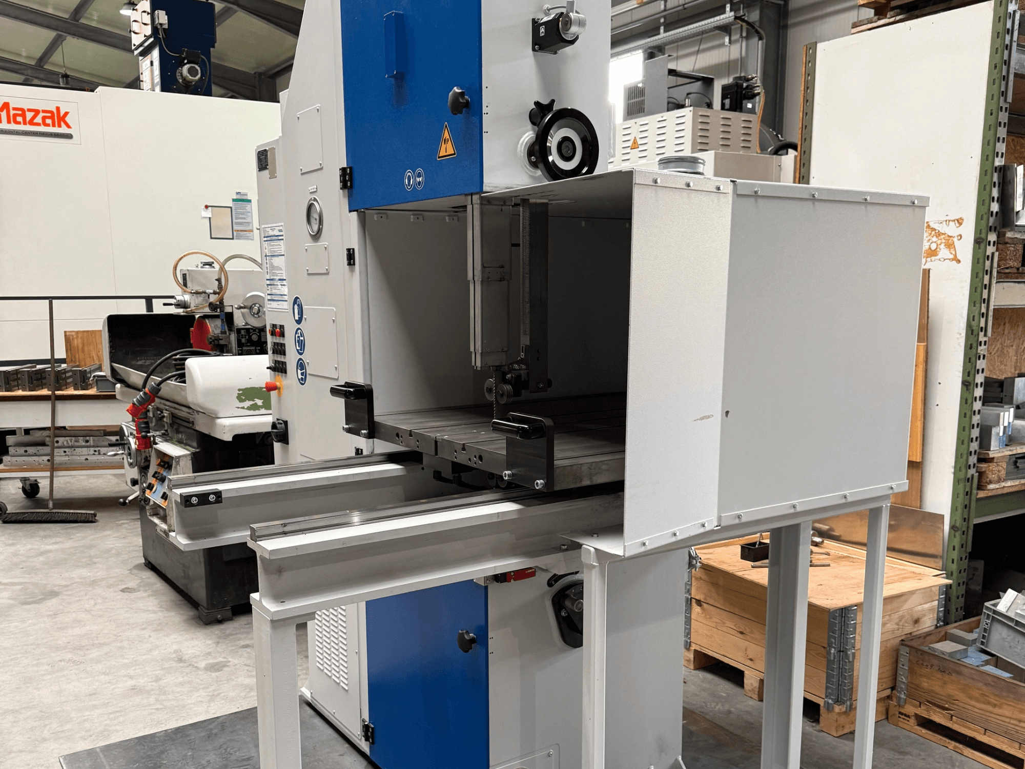 Mazaki tööstuslik töömasin esiplaanilt, millel on sinine juhtpaneel, metallist laud ja ohutushoiatused.