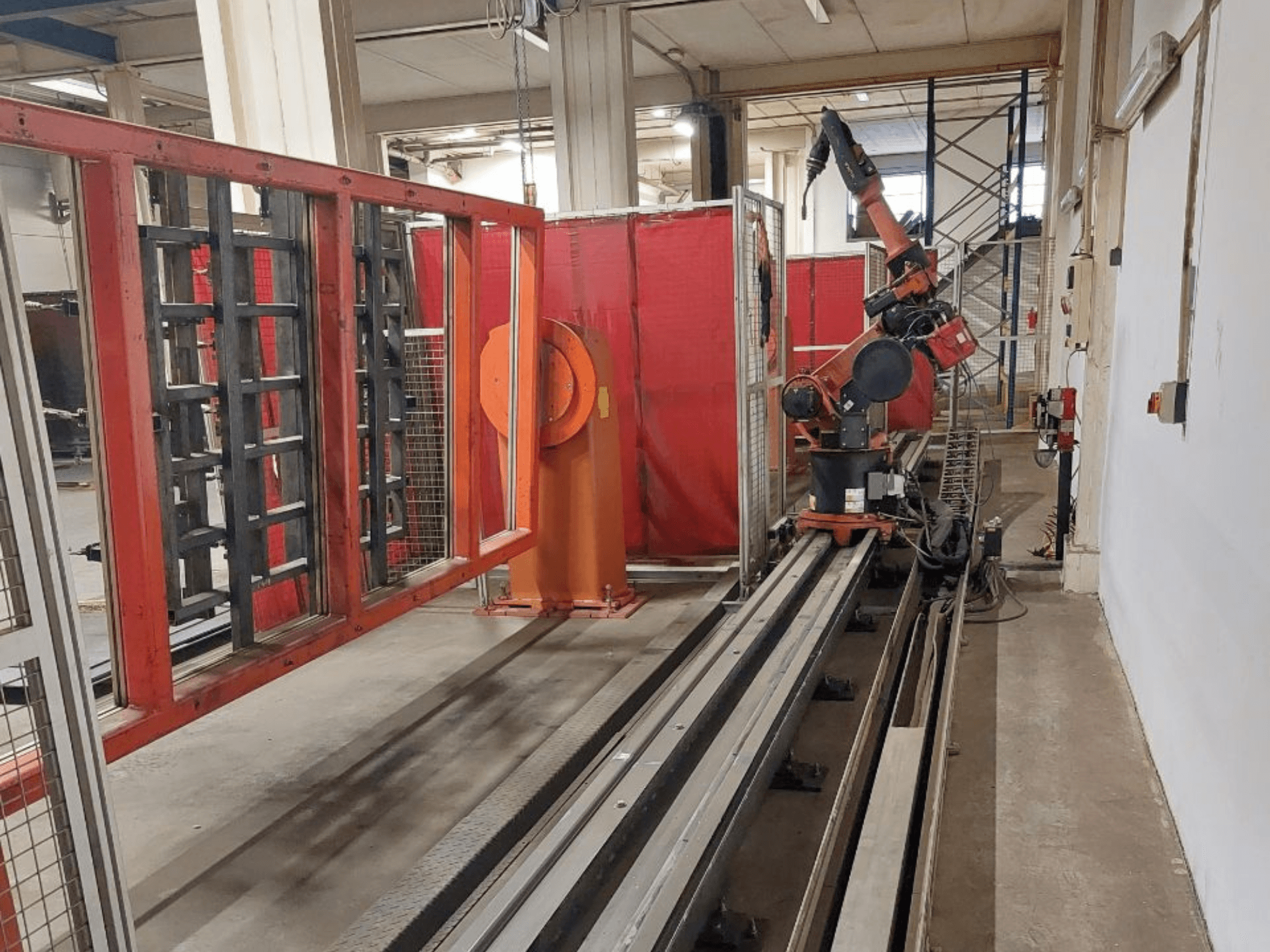 Tööstuslik oranži ja musta värvi robotkäsi, mis on paigutatud metallrajadele, ümbritsetud punaste kaitsebarjääridega, esiplaanil.