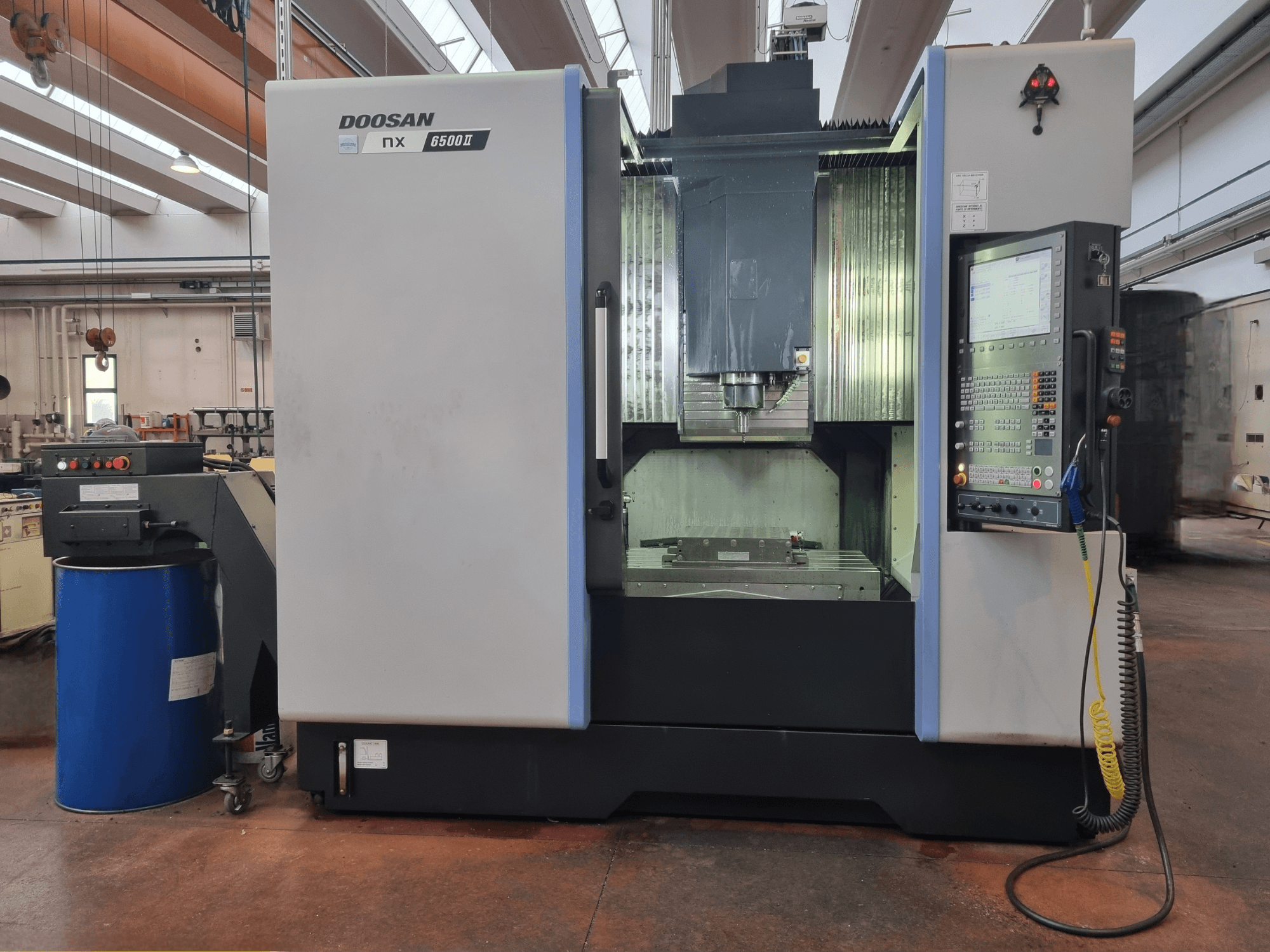Doosan NX 6500 II CNC-tööpink, esivaade, kus on juhtpaneel ja töötlemisala, mis on paigutatud sinise tünni kõrvale.