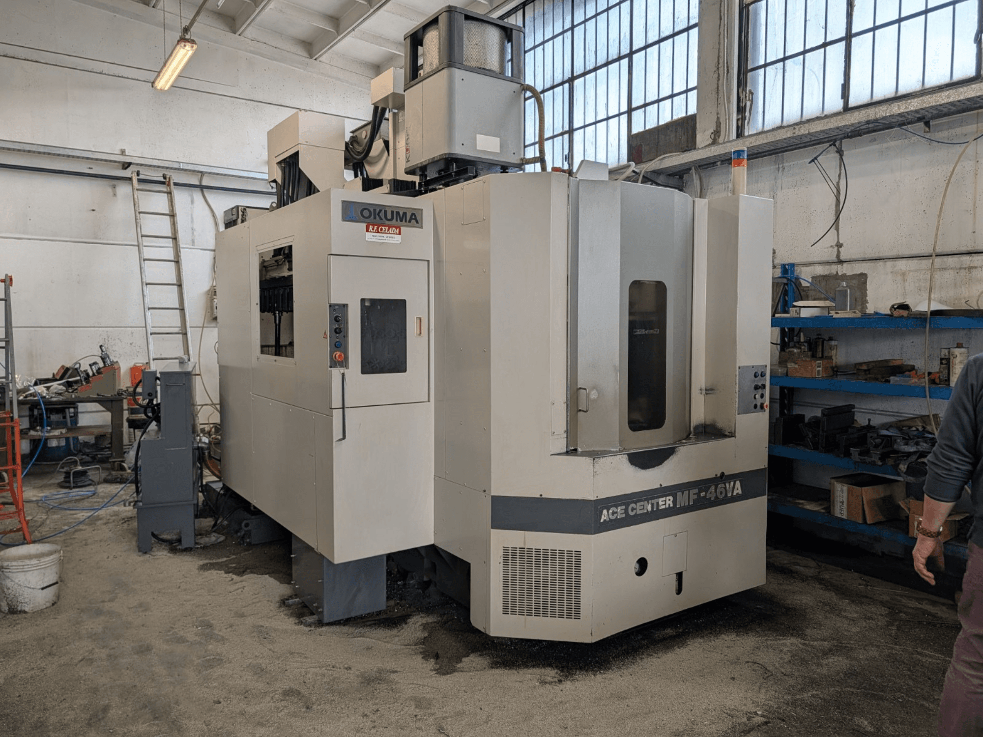Okuma ACE CENTER MF-46VA vertikaalne töötluskeskus töökojas, esiplaan koos juhtpaneeli ja ümbritsevate seadmetega.