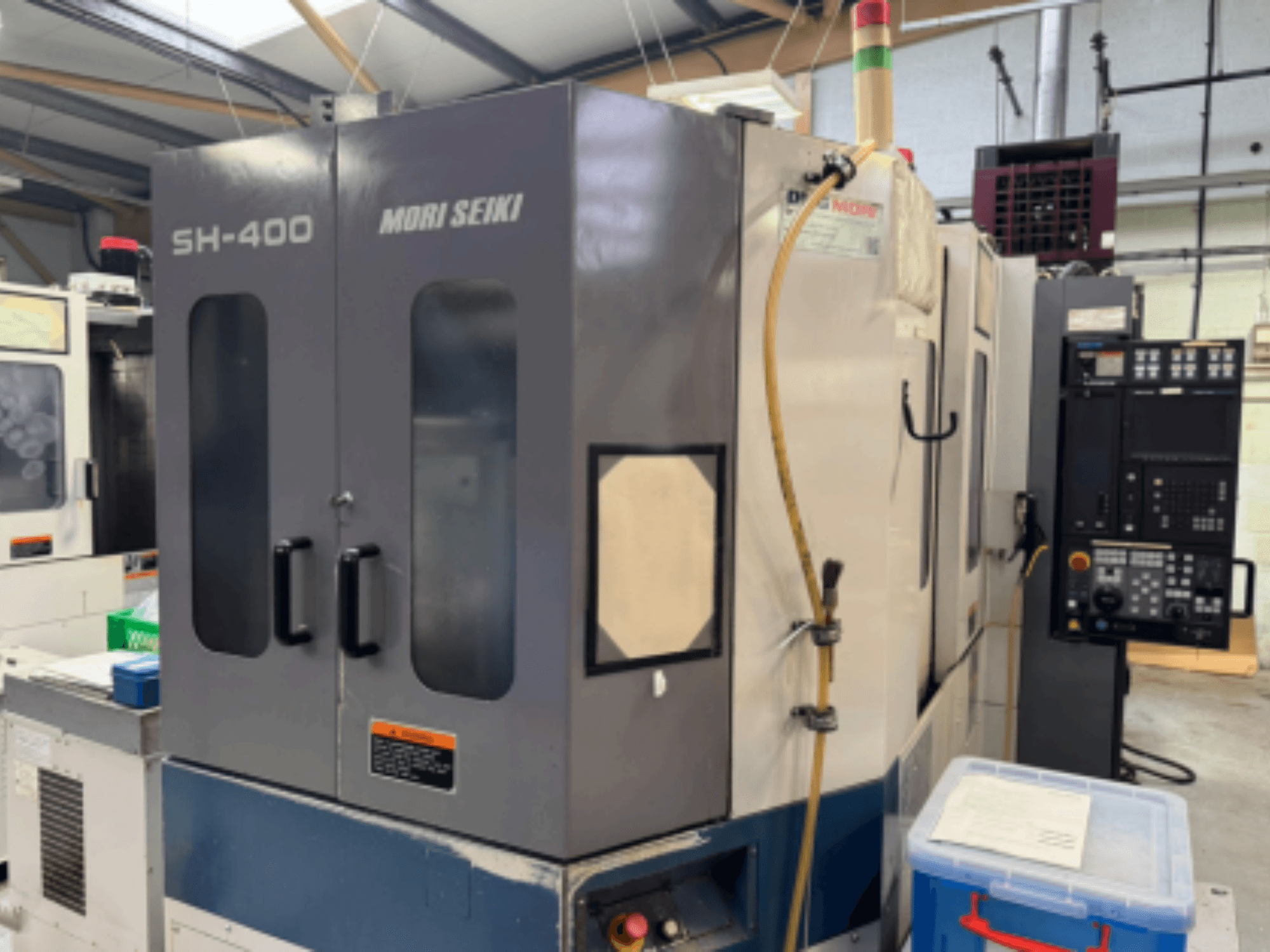 Mori Seiki SH-400 CNC-tööpink, esivaade, millel on kaks ust, juhtpaneel ja kinnitusvoolik. Lähedal sinine hoiukonteiner.