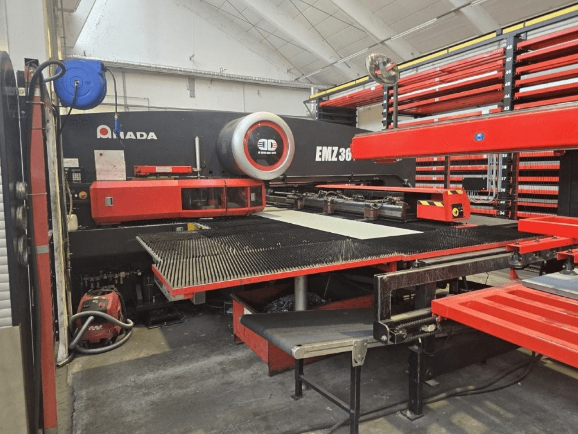 Amada EMZ 36 laserlõikepink töökojas, esiplaanil, punased aktsendid ja suur lõikelaud.