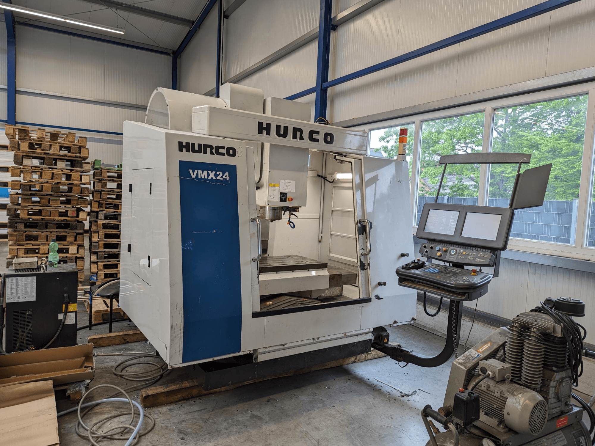 Hurco VMX24 freespink, esivaade, juhtpaneel ja tööriistade ala; taustal virnastatud kaubaalused; tööstuslik tööruum.