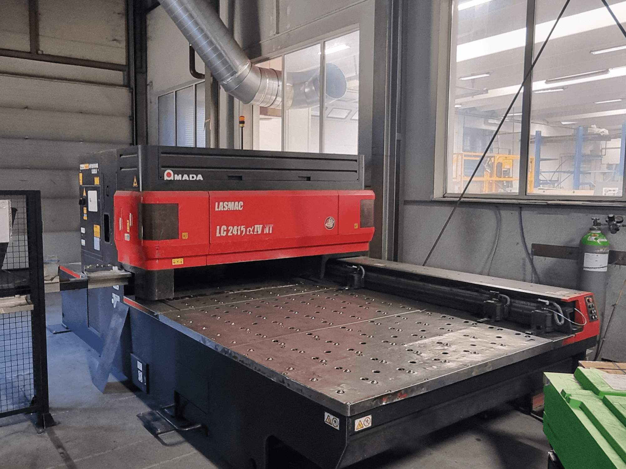 Amada LASMAC LC 2415 αⅡ NT laserlõikepink punase ja musta värviga, esiplaanil, mootoriga lõikepink ja täpsed juhtimisseadmed.