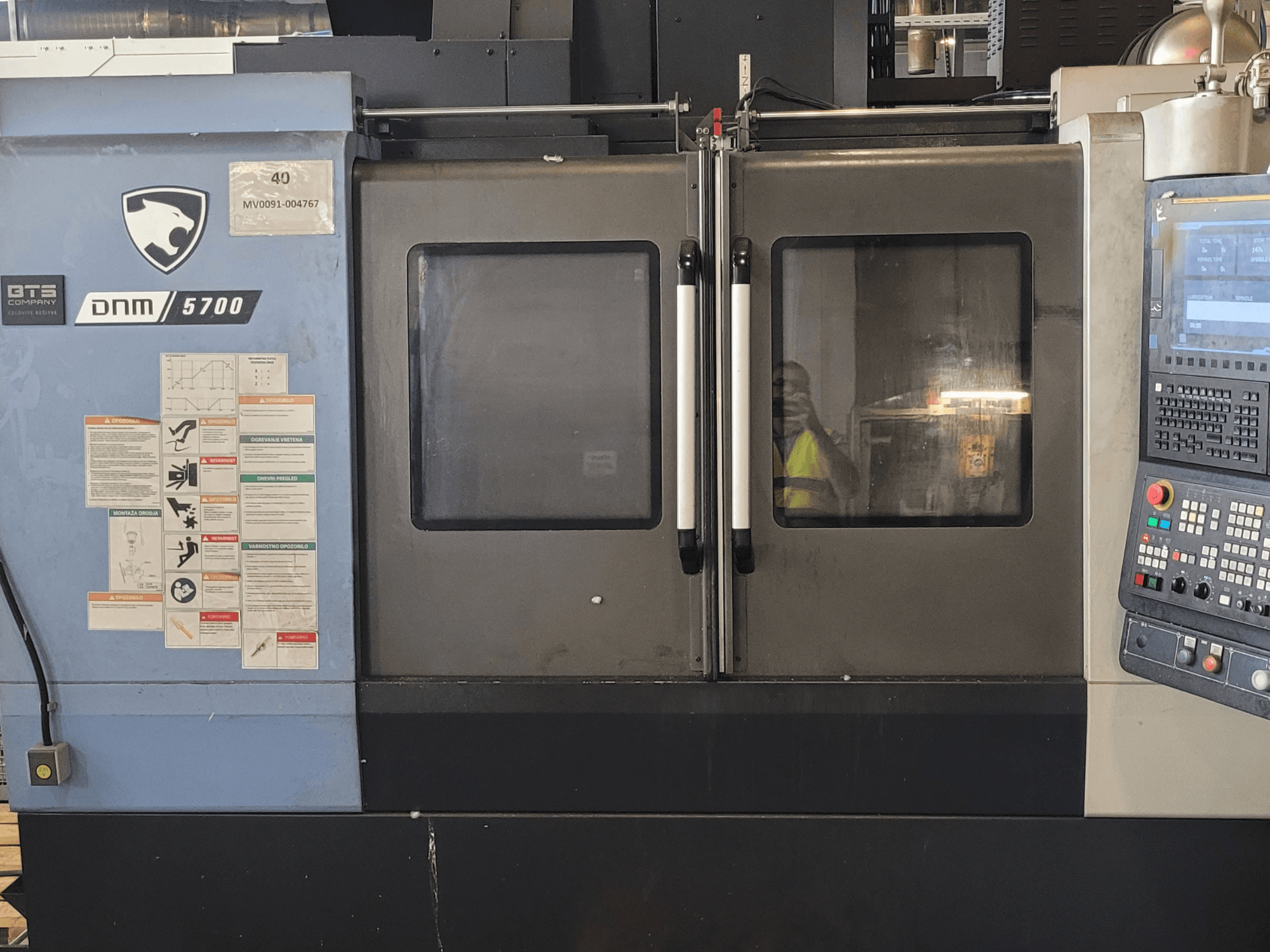DOOSAN DNM 5700 vertikaalne töötlemiskeskus, vaade eest, kus on näha juhtpaneel, ohutusmärgised ja veidi paokil olevad uksed.