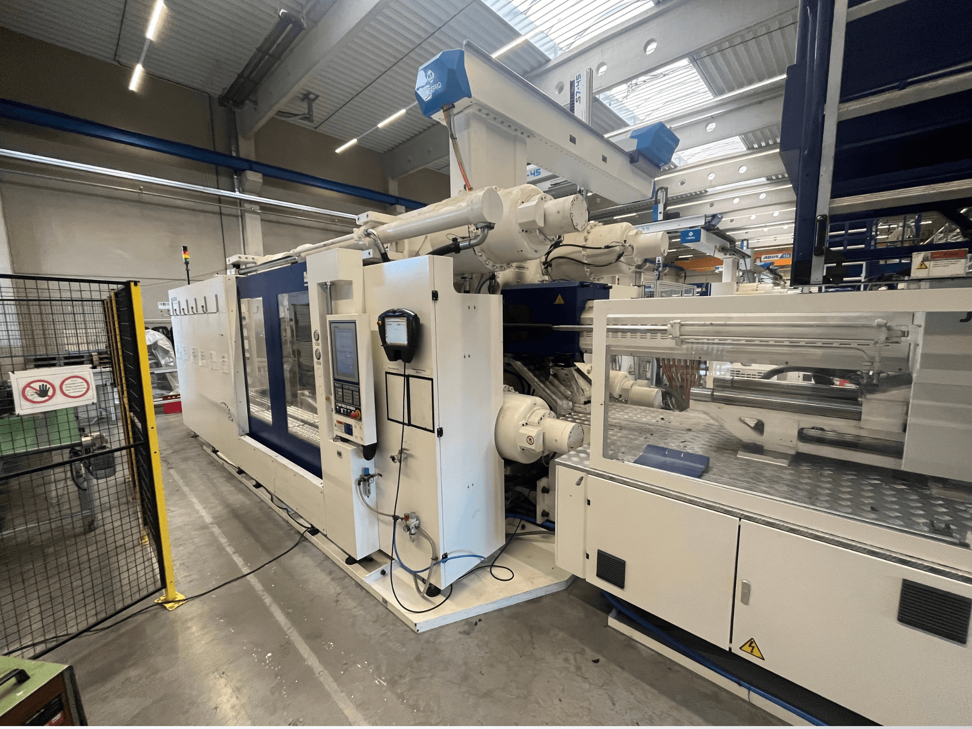 Valge ja sinise värviga survevalu masin, esivaade, millel on juhtpaneel, robotkäsi ja ohutusmärgid.