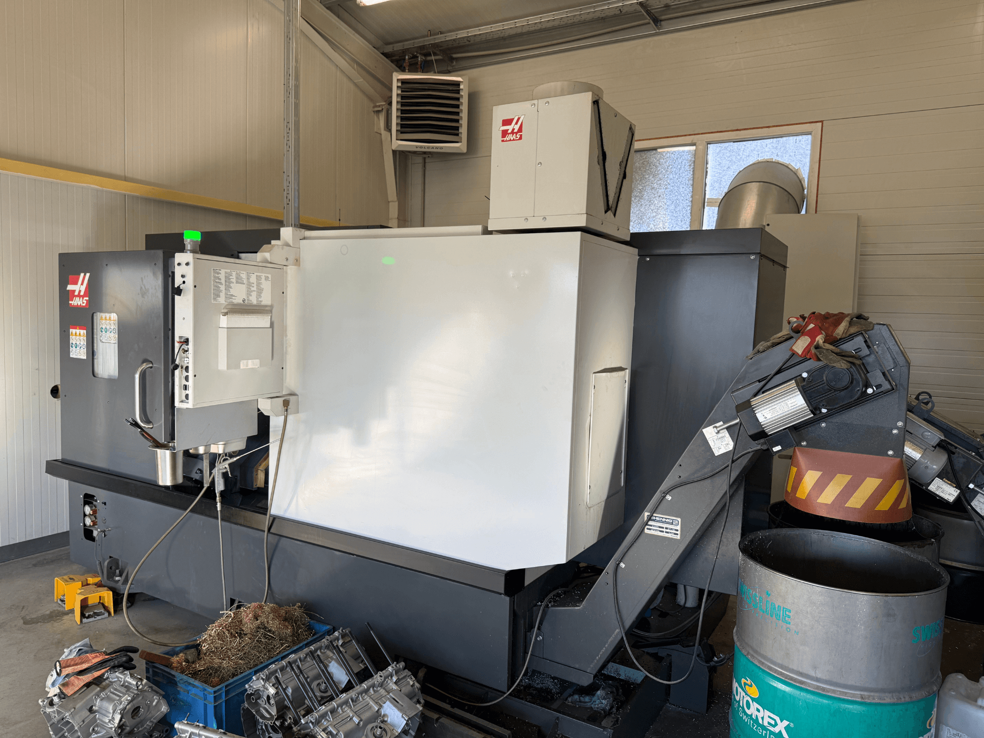 Haasi CNC-pingi juhtpaneeliga, külgvaade; nähtavad jahutusvedeliku- ja õliliinid; metallijäätmed sinises prügikastis; tööstuslik keskkond.