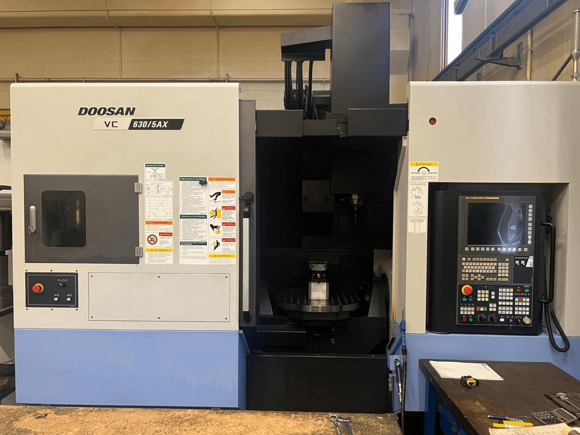 DOOSAN VC 630/5AX vertikaalse töötlemiskeskuse esivaade, millel on näha juhtpaneel ja spindli piirkond koos nähtavate ohutusmärgistega.