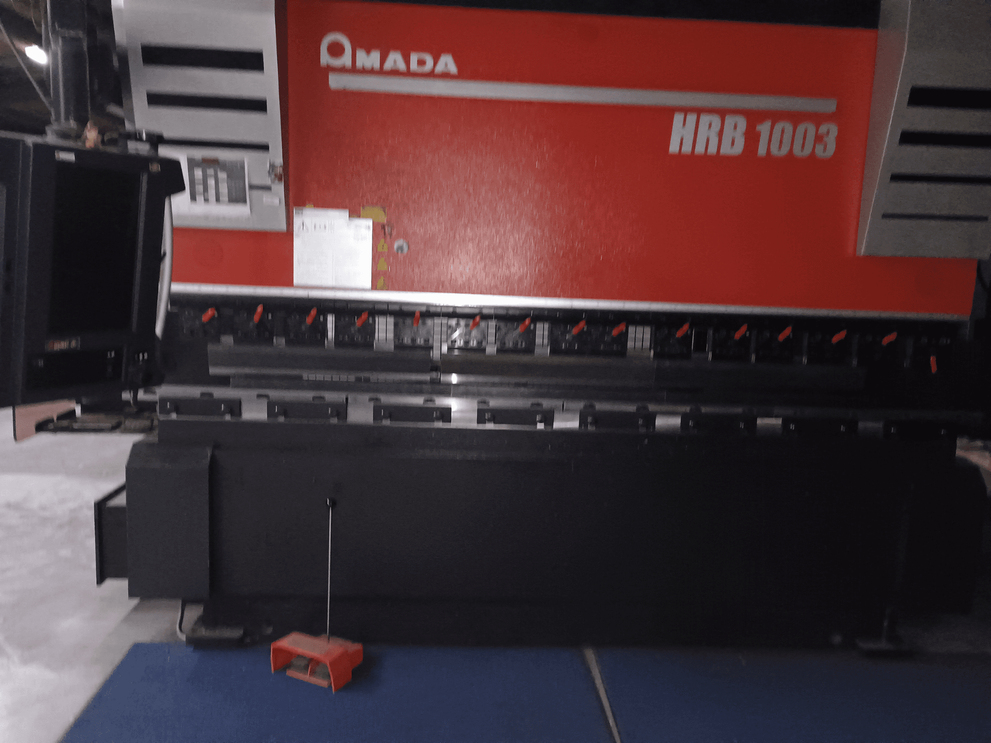 Amada HRB 1003 pressipidur, esiplaan; punaste nuppude ja musta korpusega juhtpaneel, mis on paigutatud tööstuskeskkonda.