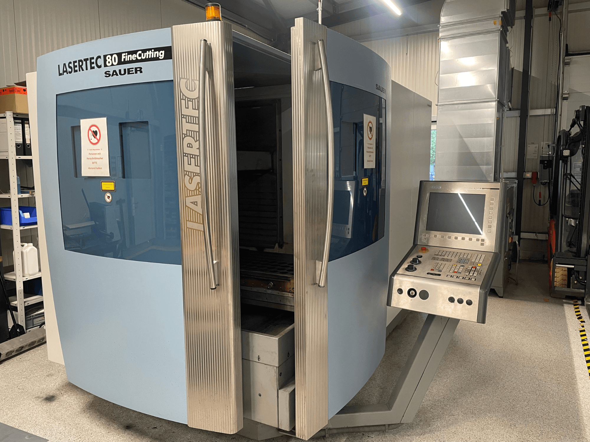 DMG Sauer Lasertec80 FineCutting lehtmetalli töötlemismasina esivaade koos juhtpaneeliga, avatud ustega ja nähtavate ohutusmärkidega.
