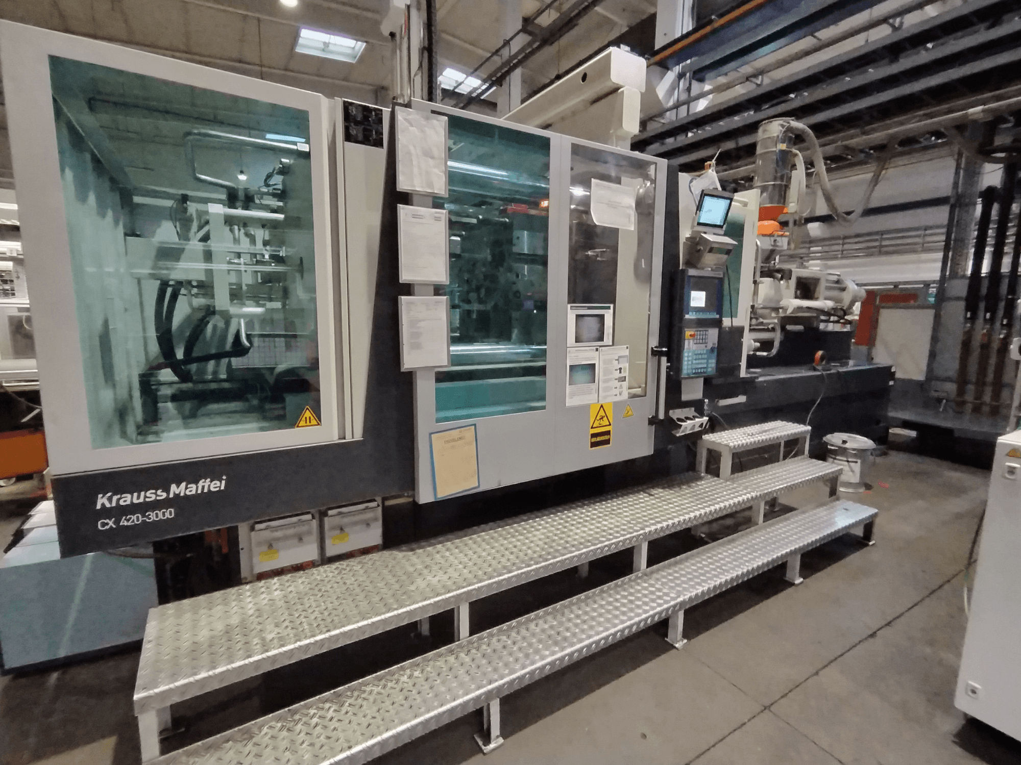 Krauss Maffei CX 420-3000 survevalu masin, külgvaade, juhtpaneel ja ohutussildid nähtavad, metallist treppidel.