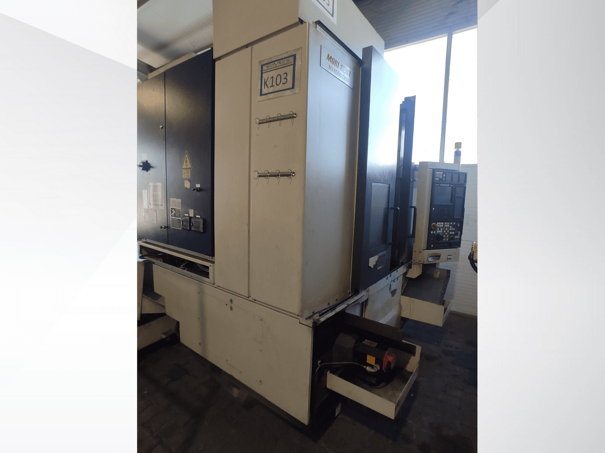 Mori Seiki K103 CNC-tööpink eesvaates, kus on näha juhtpaneel, ohutussildid ja elektrilised komponendid.