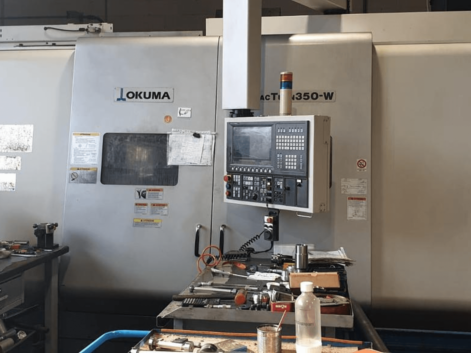 Okuma MAC TURN 350-W treipingi keskus, esivaade juhtpaneeli ja tööriistade ja pudeliga täidetud tööruumiga.