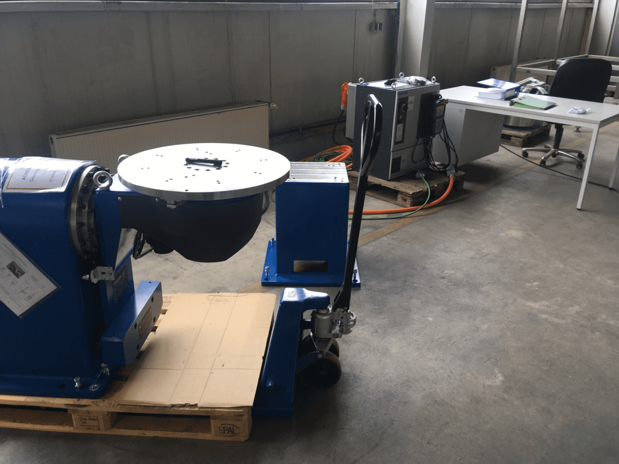 Sinine tööstusseade ümmarguse aluse ja juhtpaneeliga, esiplaanilt vaadatuna, puidust kaubaalusel kontoris.