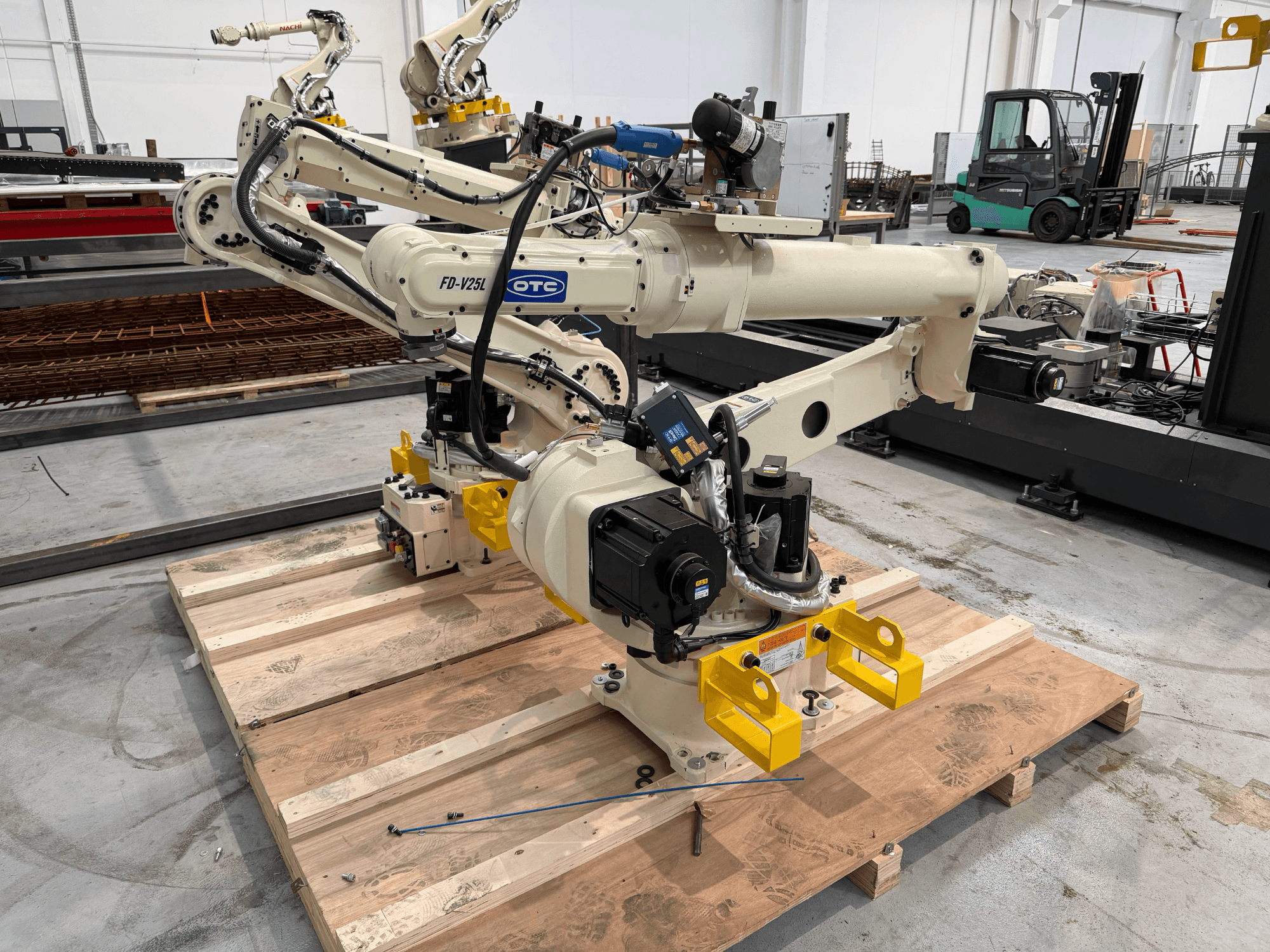 OTC Daiheni robotkäsi mudel FD19-V25L vaates eest, millel on kollased haardeküünised ja nähtavad mehaanilised osad puidust kaubaalusel.