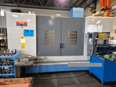 Mazak VTC-300C masin tehases, esiplaanil on juhtpaneel, hallid uksed koos aknaruudustikuga ja lähedalasuvad seadmed.