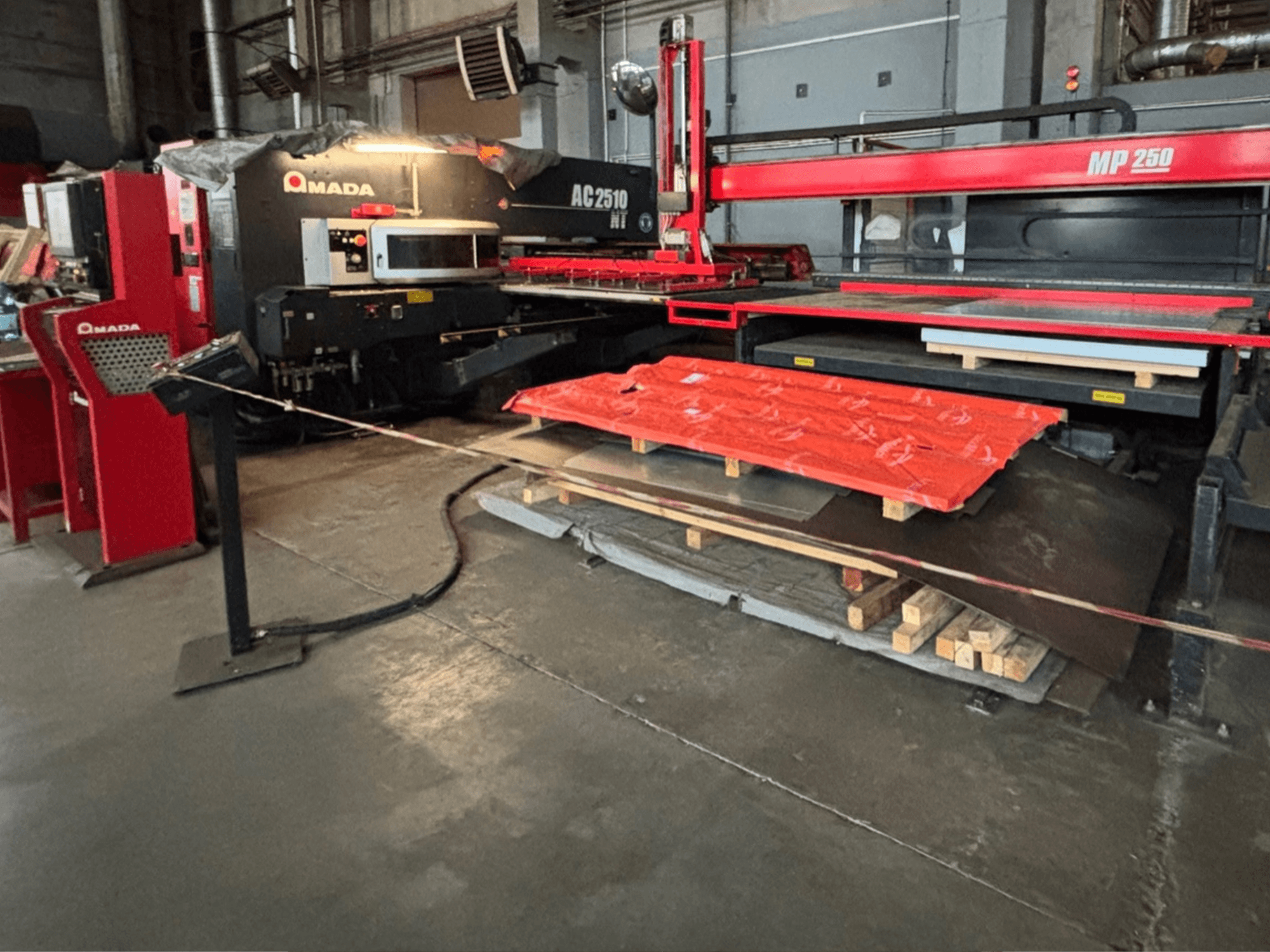 Amada AC2510 NT laserlõikur tehases, esiplaanil, raske masin koos metallplaatide ja kaubaalustega põrandal.