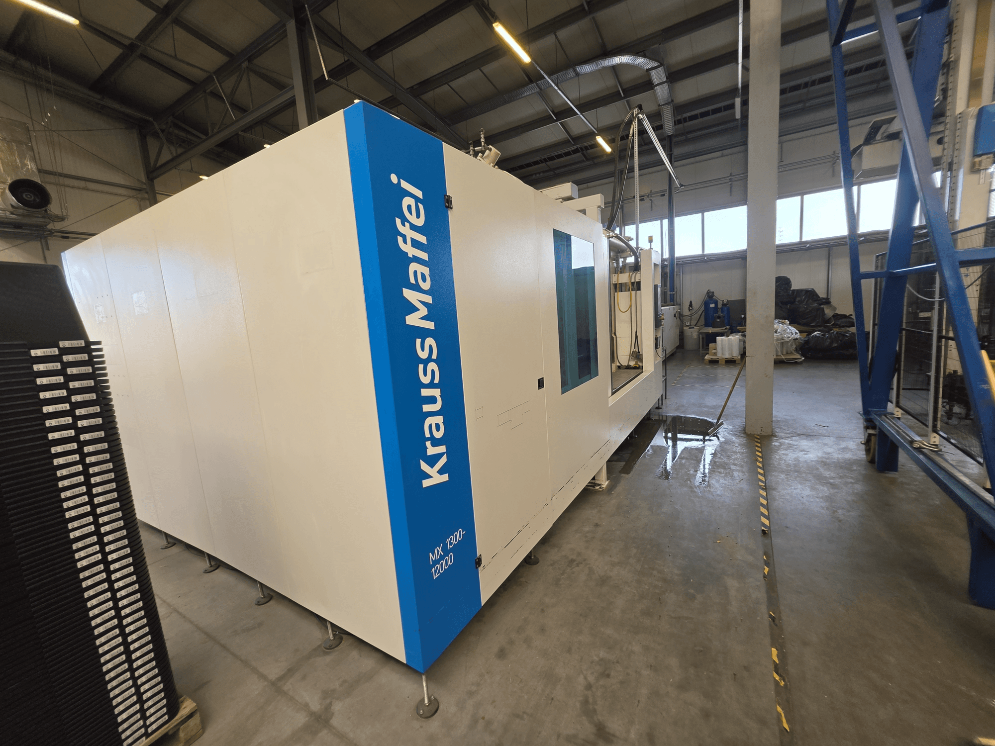Krauss Maffei MX 1300-12000 hüdrauliline süstimisvormimismasin külgvaates, millel on näha suur valge šassii ja sinine logoga märgistus.