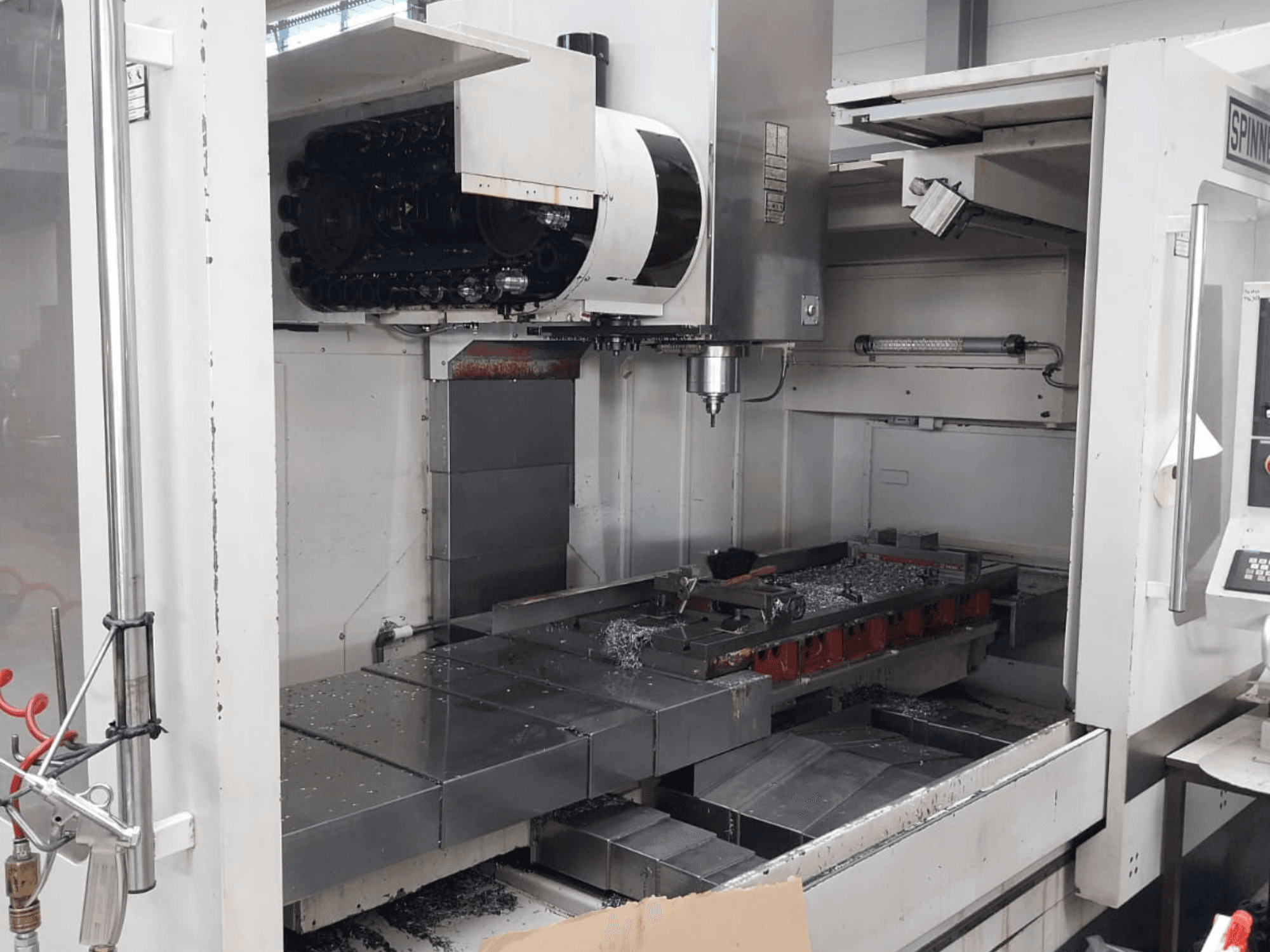 SPINNER MVC 1600 vertikaaltöötluskeskuse esiplaan, kus on näha avatud tööruum, spindel ja metallilaastud laual.