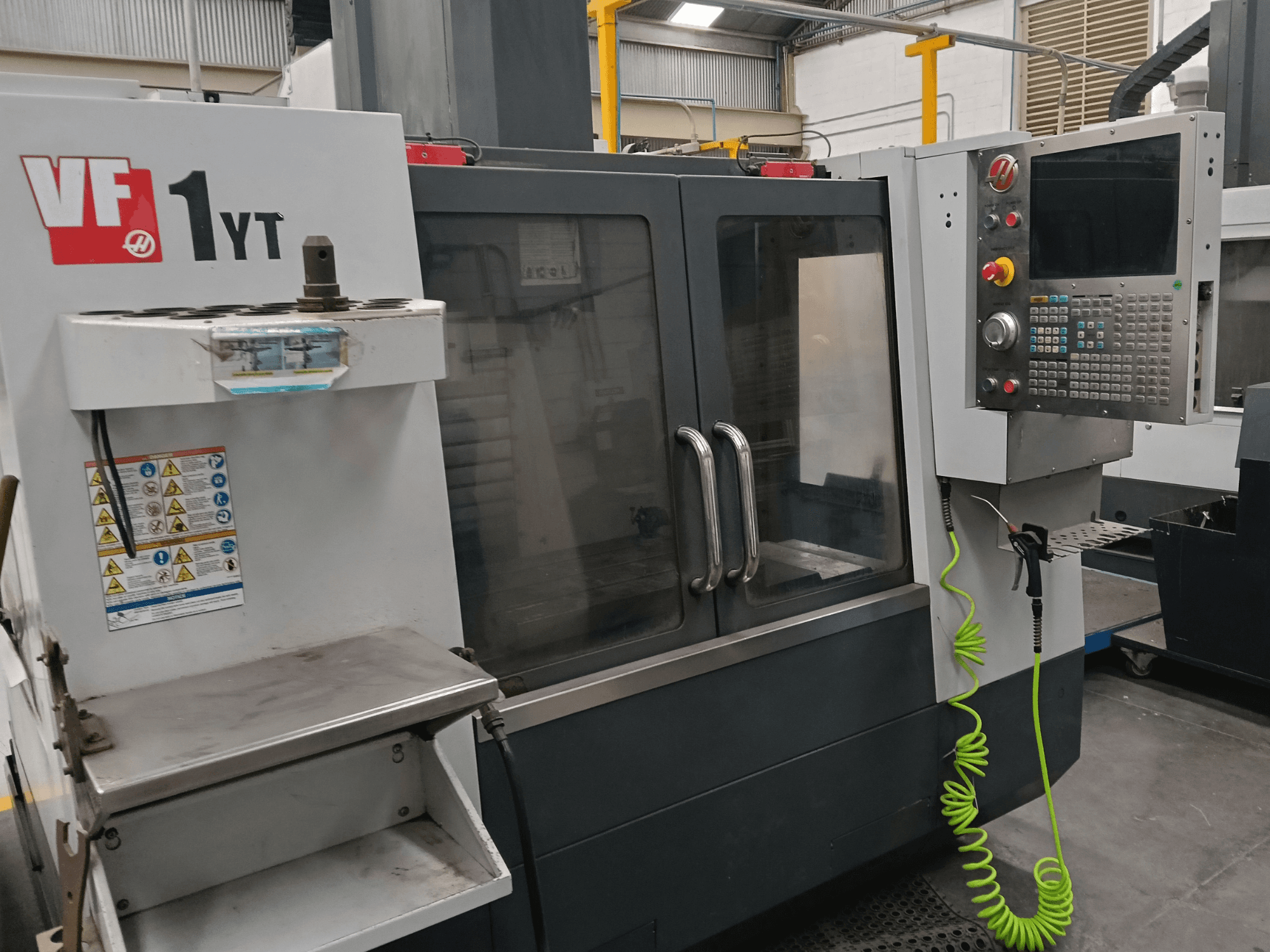 HAAS VF-1YT-2 vertikaalne töötlemiskeskus, vaade eest, millel on näha juhtpaneel, tööriistahoidja, ohutusmärgised ja töötlemisala.