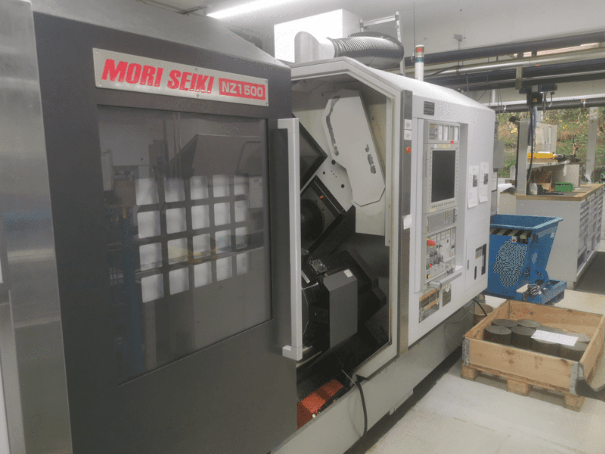 MORI SEIKI NZ1500 CNC-tööpink kerge nurga all, kus on näidatud juhtpaneel ja tööruum koos metallitoorikutega puidust kaubaalusel.