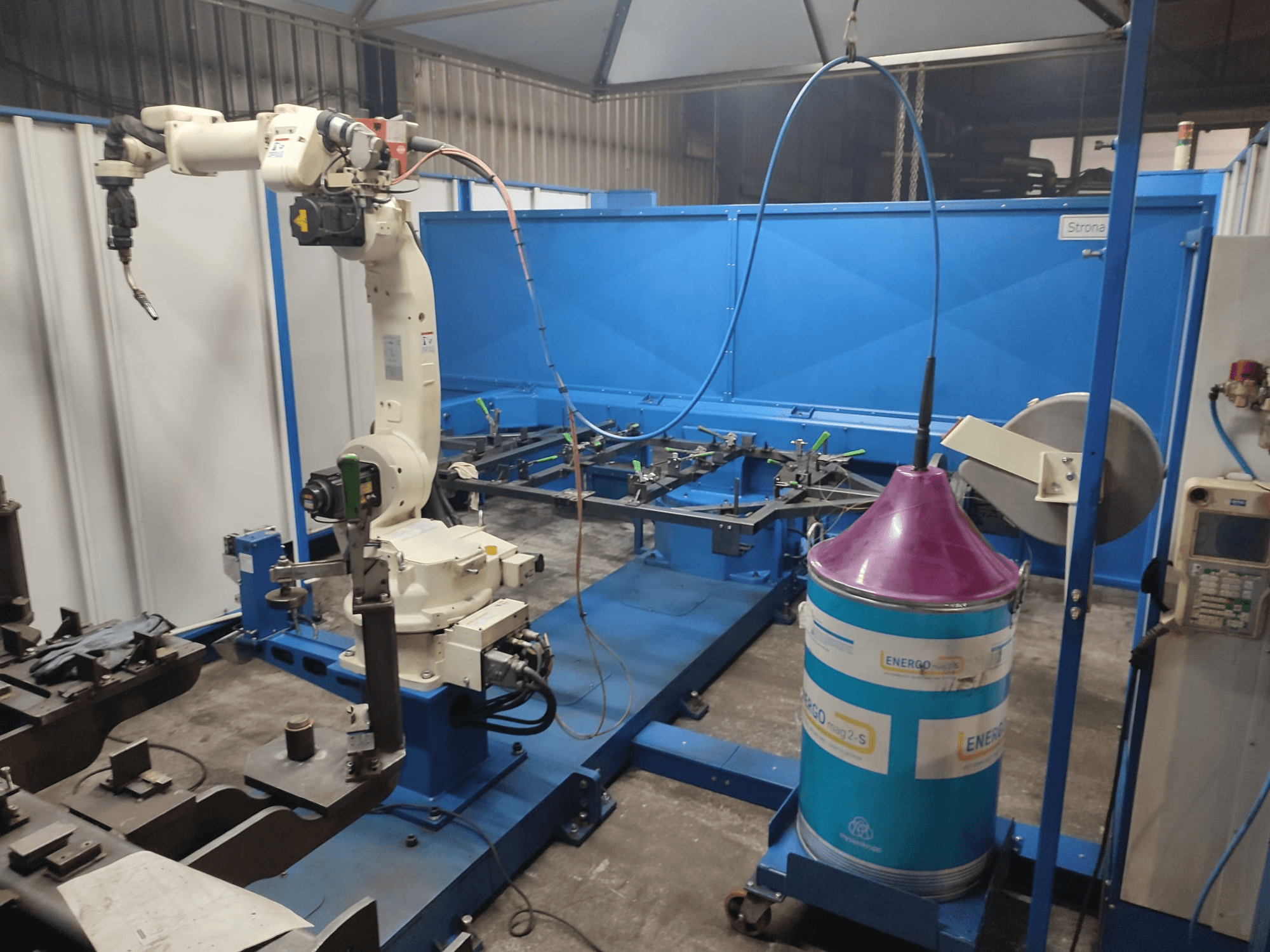 Valge ja sinise alusega tööstuslik robotkäsi, mis on seadistatud töökojas sinise tööpinnaga ja metallist komponentidega.