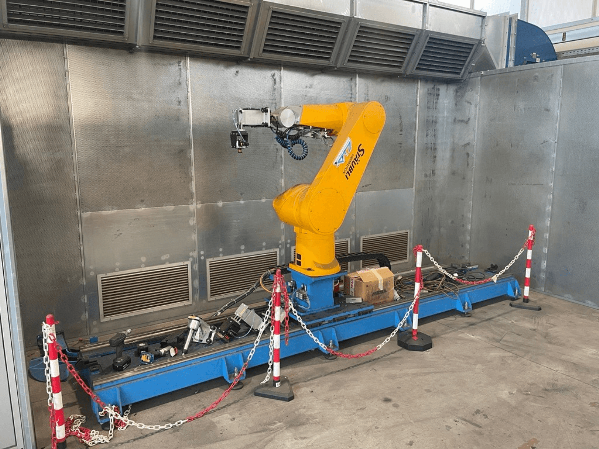 STAUBLI CS8C-RX160 robotkäsi töökojas, külgvaade, koos kaablite ja erinevate tööriistadega sinisel alusel, mida ümbritsevad kaitsepiirded.