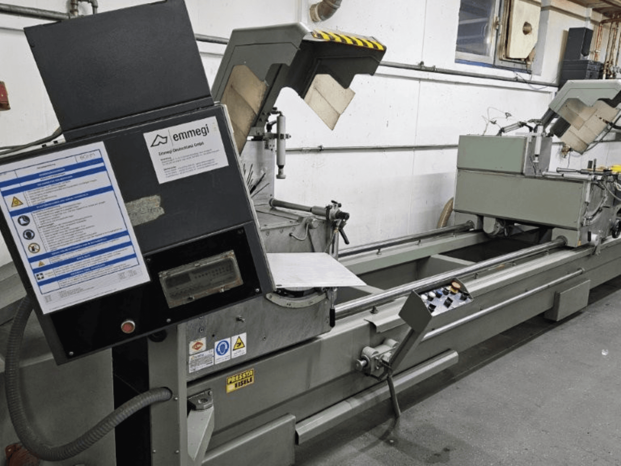 Emmegi tööstuslik lõikemasin, esivaade, millel on juhtpaneel, ohutussildid ja töökomponendid.