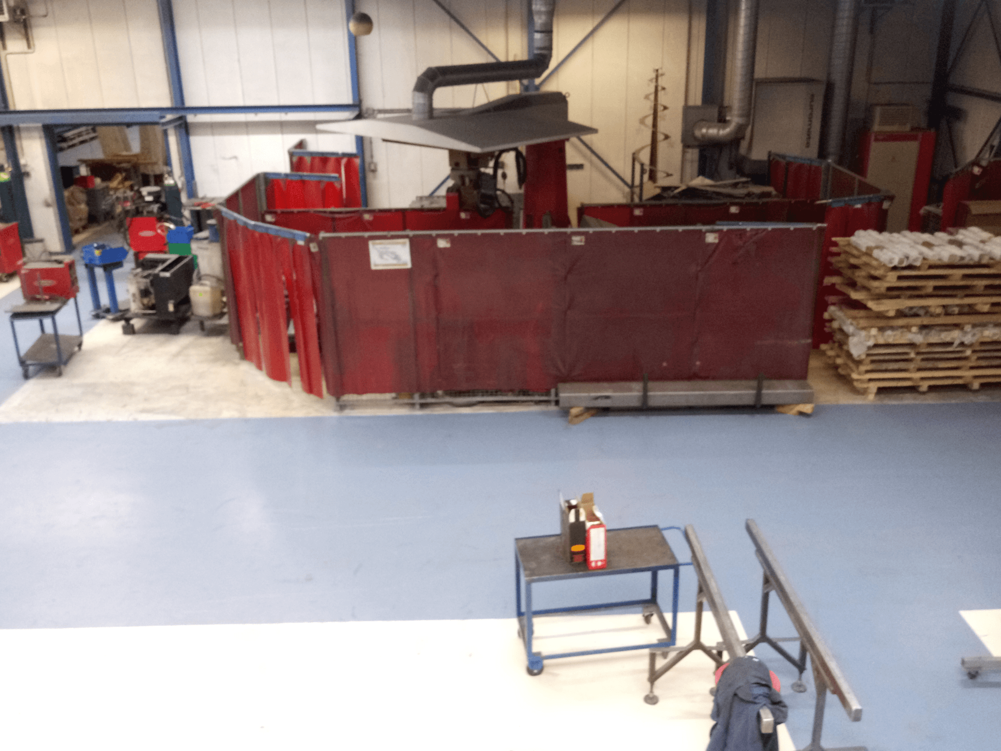 Tööstuslik tööruum, kus on punased keevituskardinad, masinad ja kaubaalused; ülaltvaade, mis näitab paigutust ja seadmeid.