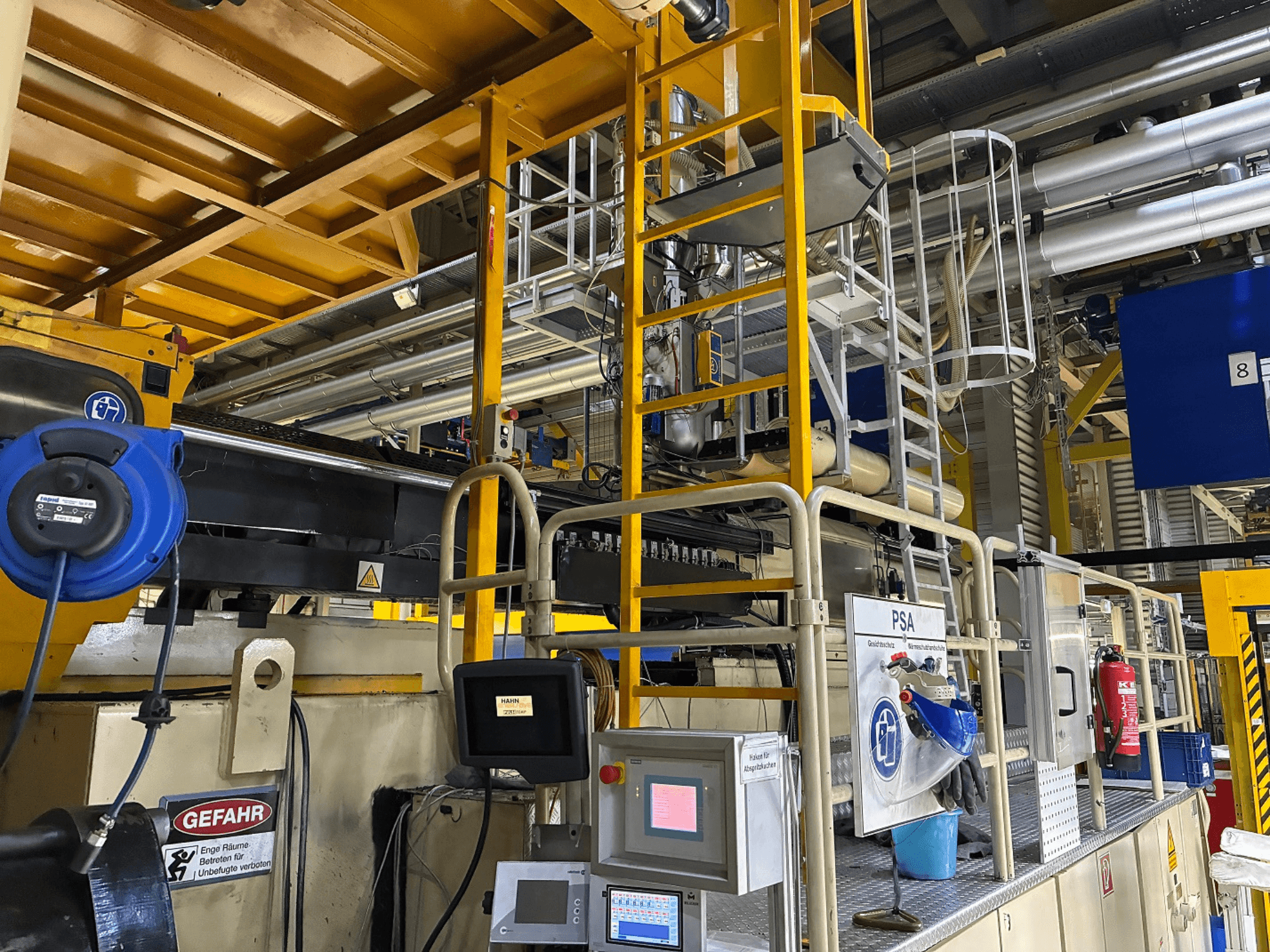 Tööstusmasinate juhtpaneel siniste ohutusseadmete ja kollaste tellingutega, külgvaates on näha torud ja hoiatusmärgid.