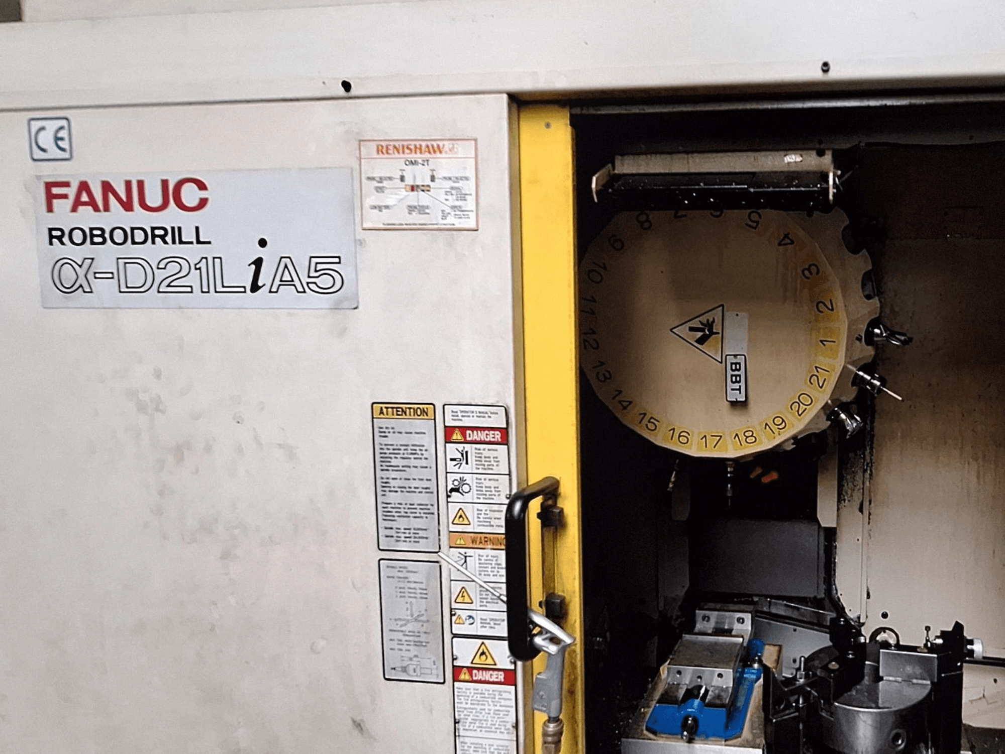 FANUC Robodrill α-D21LiA5, esiplaanil. Näitab tööriistakarusselli ja töötlemisala koos klambritega ning hoiatusi uksel.