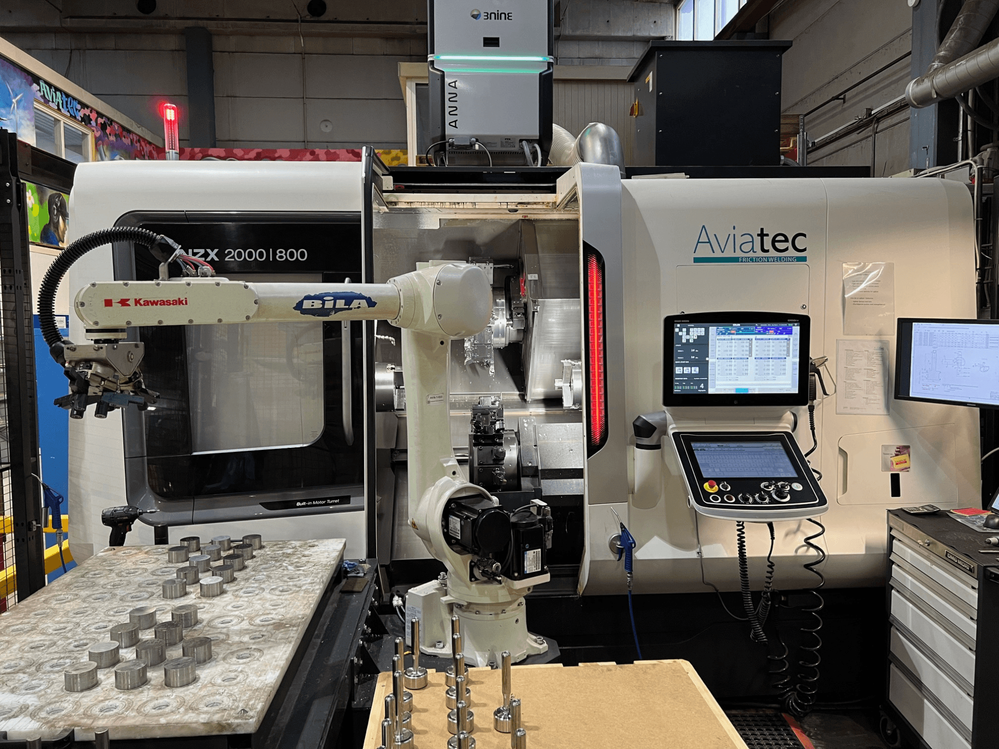 Horisontaalne treipink DMG MORI NZX 2000/800SY koos Kawasaki robotkäega, juhtpaneeliga ja tööpinnal asuvate metallosadega.