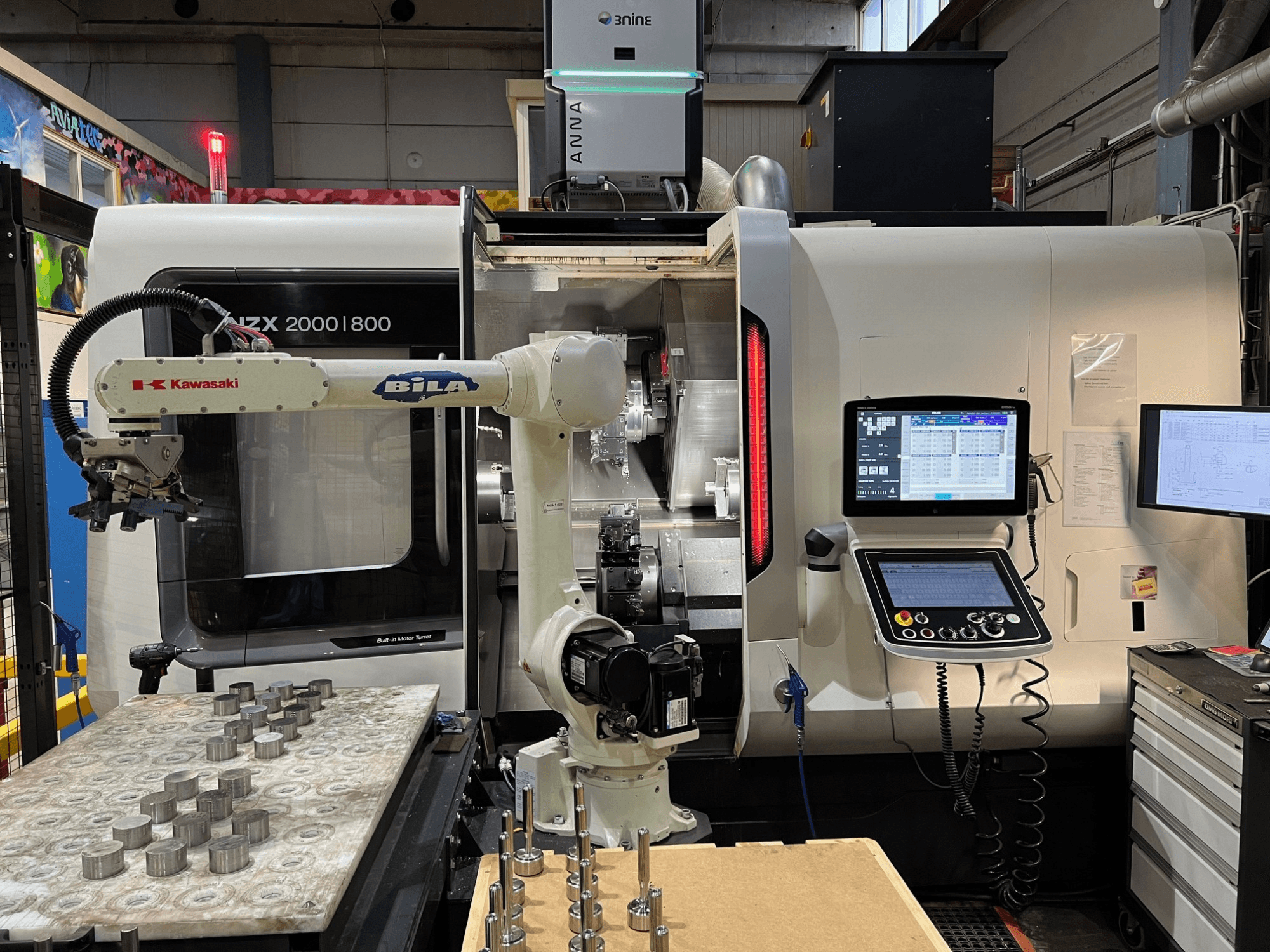 Töökojas asuv horisontaalne treipink DMG MORI NZX 2000/800SY, millel on näha Kawasaki robotkäsi ja juhtimispaneelid.