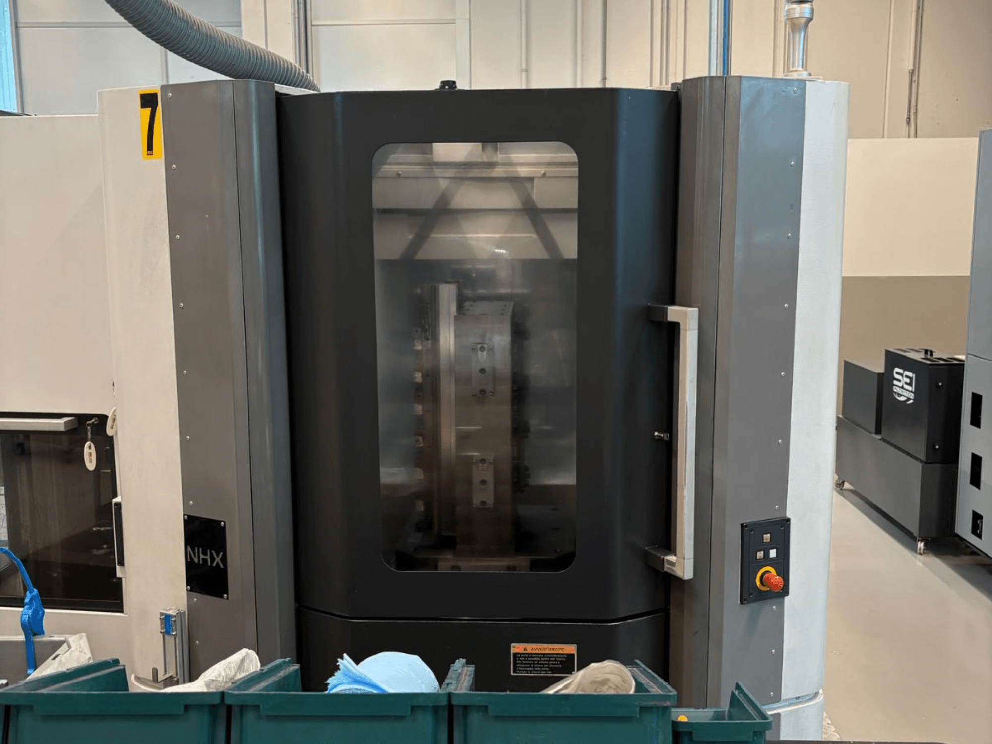NHX CNC-tööpink eesvaates, kus on näidatud juhtpaneel ja selge ekraan; asub töökojas, mille lähedal on rohelised prügikastid.