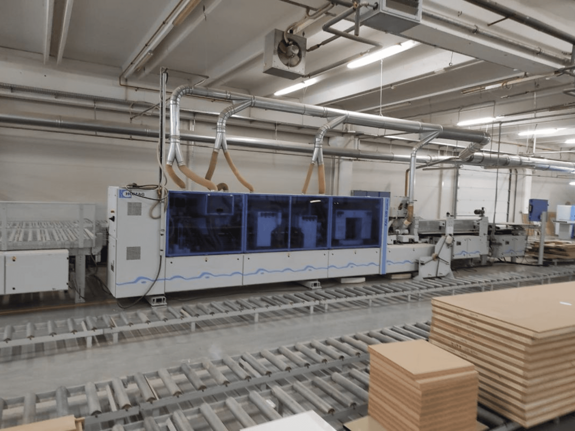 HOMAGi tootmismasin tehases, esiplaanil, sinise korpuse ja konveierisüsteemiga, millel on virnastatud vineerplaadid.