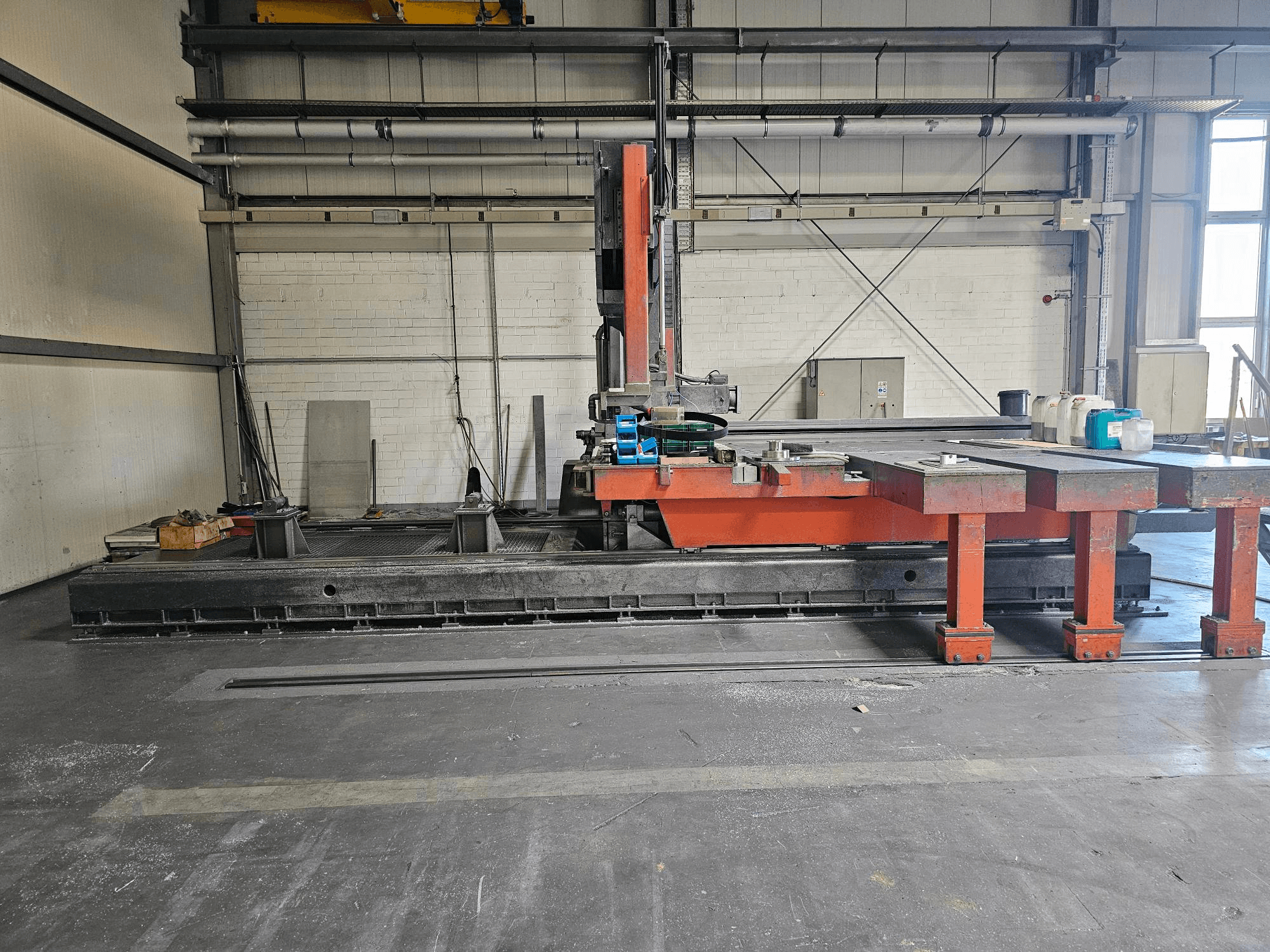 Tööstuslik lõikepink avatud töökojas, oranž ja must, suure lauaga; külgvaates on näha tarvikuid ja tööruumi.