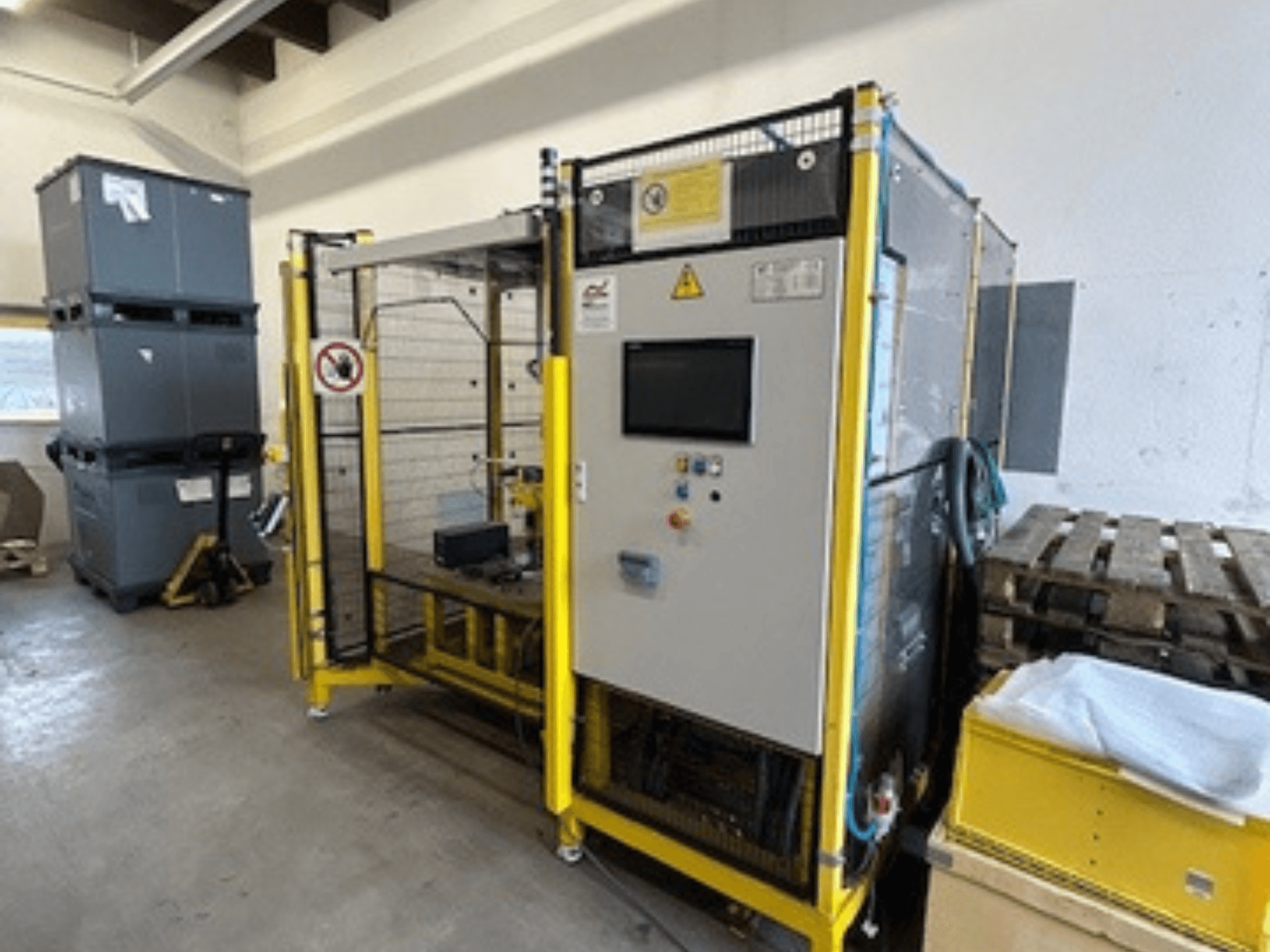 Tööstusmasin juhtpaneeliga ja ohutusseadmetega, esiplaanil; taustal kollane raam, hallid kastid ja kaubaalused.