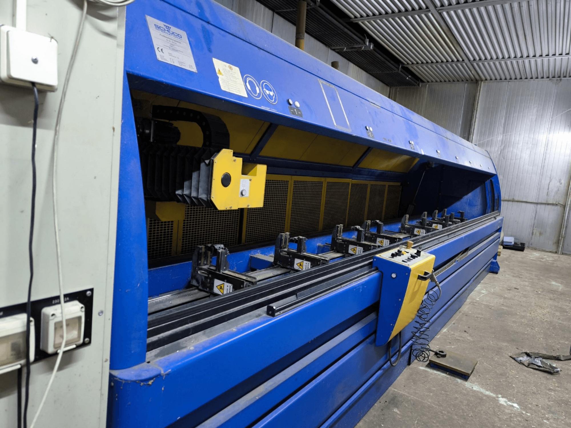Schneideri tööstuslik lõikemasin sinises toonis, külgvaates, juhtpaneel, rihmad ja mitu kinnitusjaama nähtavad.