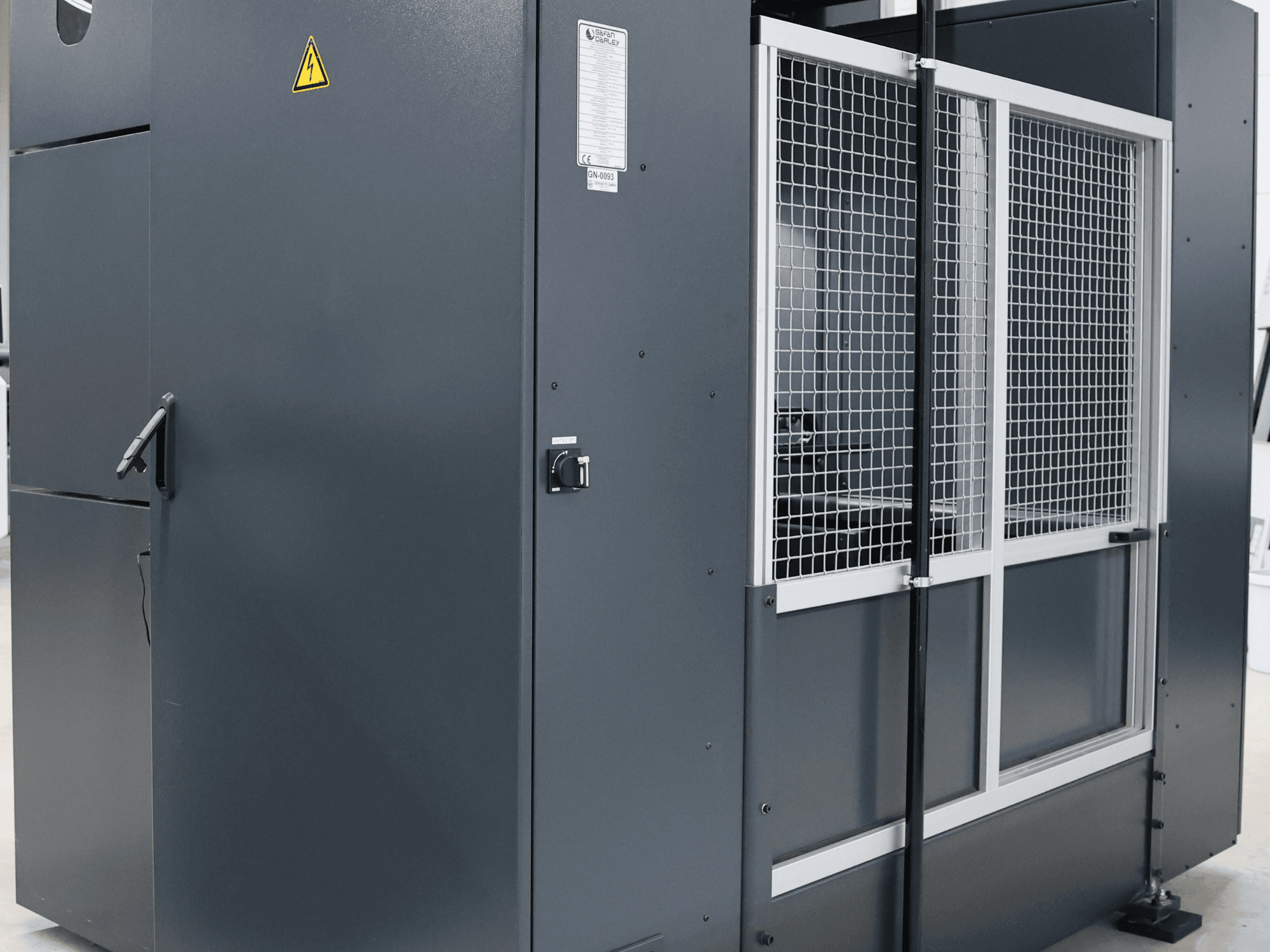 Hall tööstuslik masin ohutushoiatusega, esiplaanil võrkudega uks ja juhtpaneel, mudel on märgistatud.