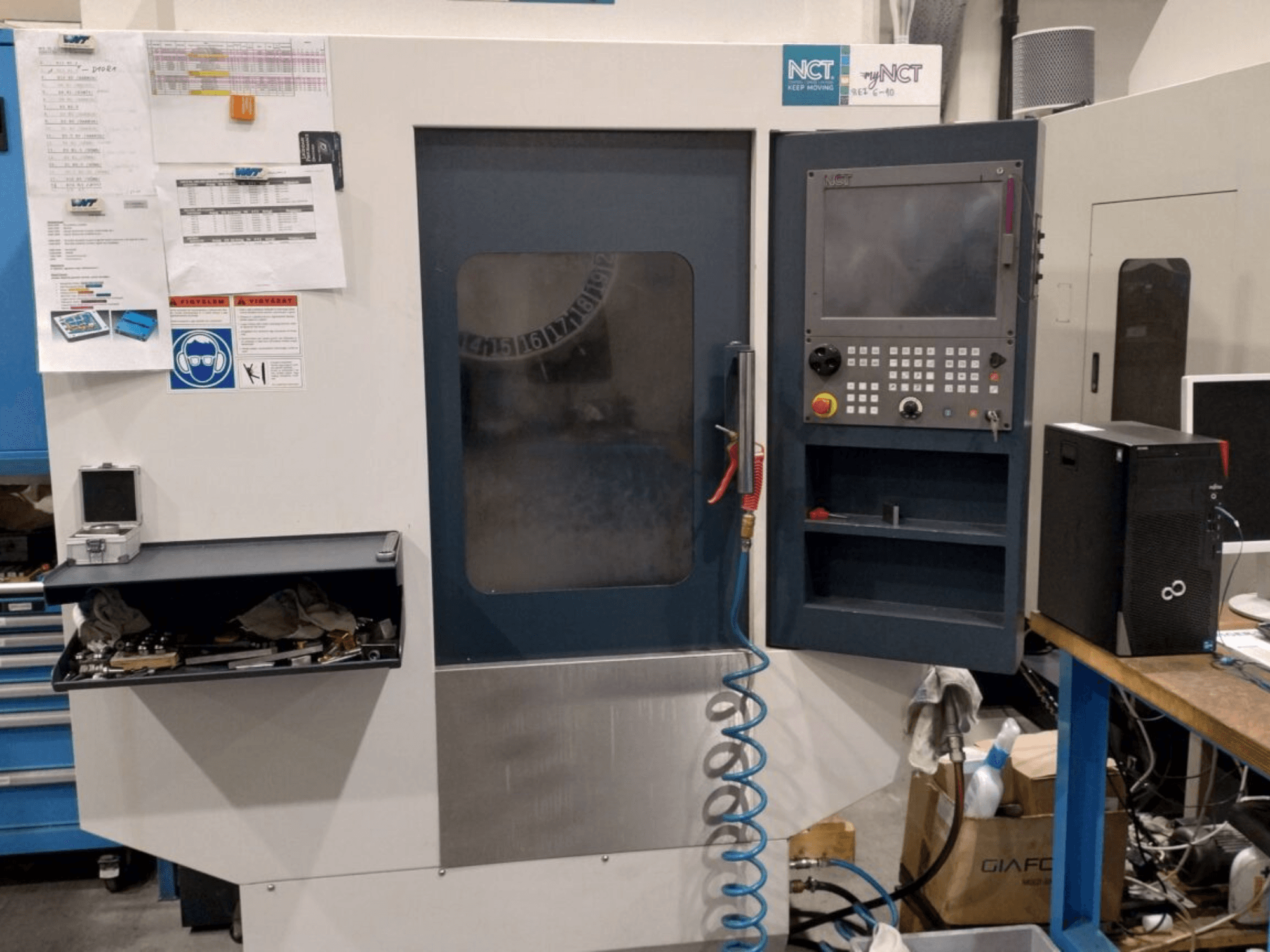 NCT CNC-tööpink, esiplaanil, juhtpaneel numbrilise klaviatuuriga, tööriistad hoiusahtlis ja käsiraamat seinal.