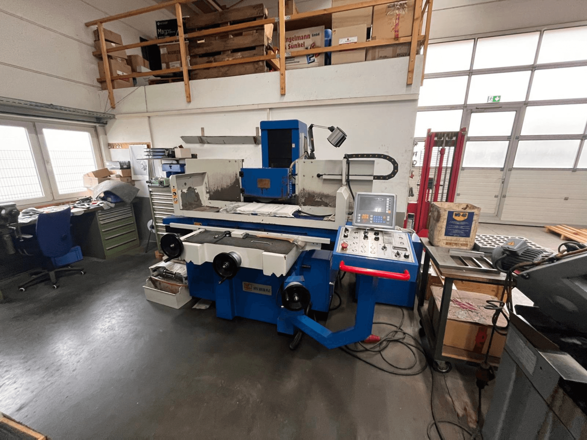 Sinine pindade lihvimismasin digitaalse juhtpaneeliga, esivaade, millel on näha tööruumi segadust ja rohelist tööriistakappi.