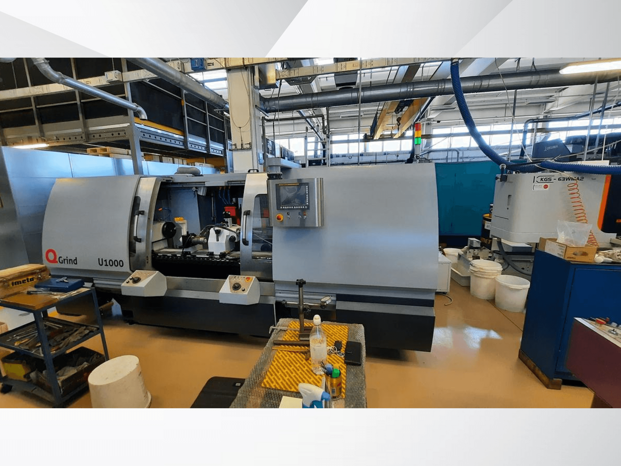 Q-Grind U1000 masin töökojas, esiplaanil on juhtpaneel ja avatud lõikepind, mida ümbritsevad seadmed.
