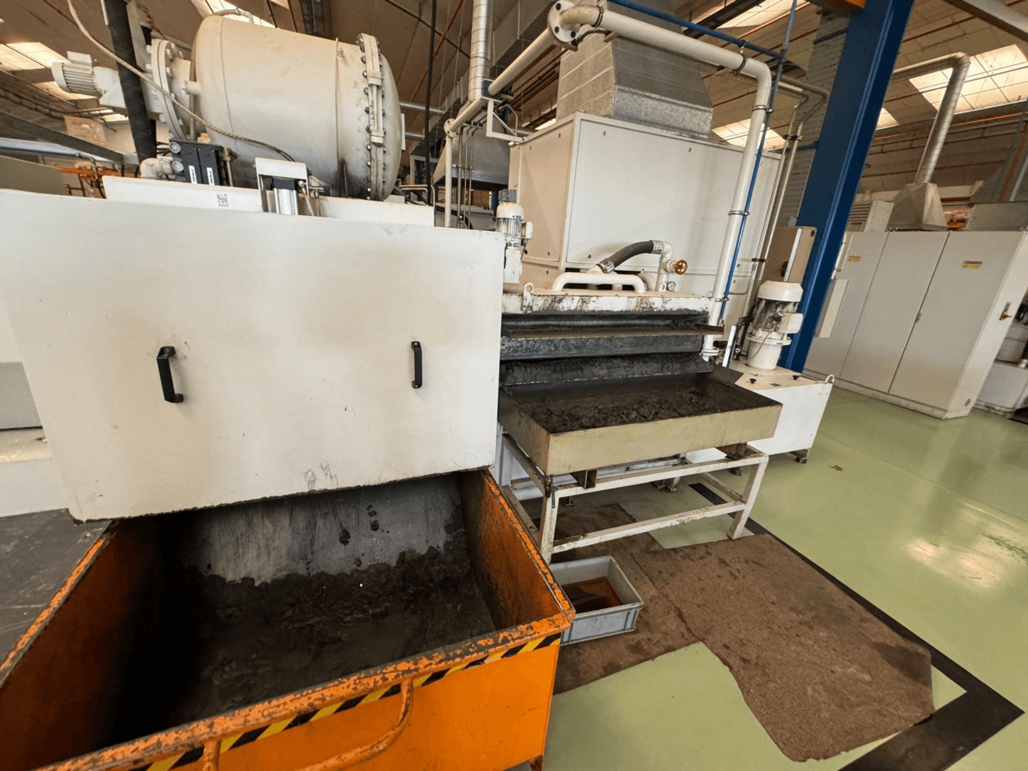 Tööstuslik töötlemisseade koos oranži prügikastiga, esiplaanil; sisaldab mootorit, konveierid ja hooldusala.