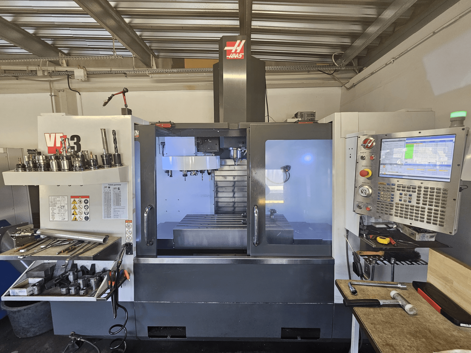 Haas VF-3 CNC-tööpink töökojas, esiplaanil tööriistade, juhtimisseadmete ja freesimisala valgustatud sisemusega.