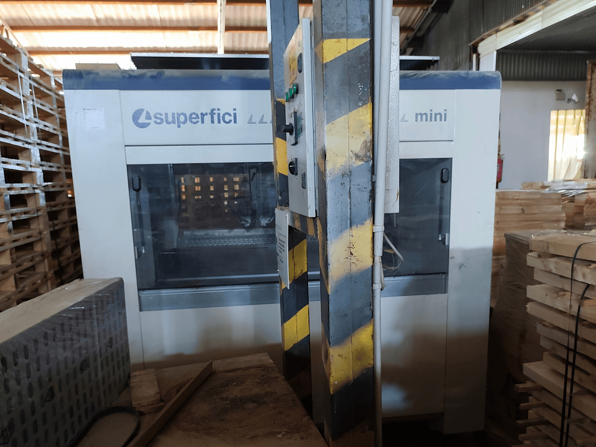 SCM Superfici Mini puiduviimistlusmasin eestvaates, millel on näha juhtpaneel ja seda ümbritsevad puidust kaubaalused.
