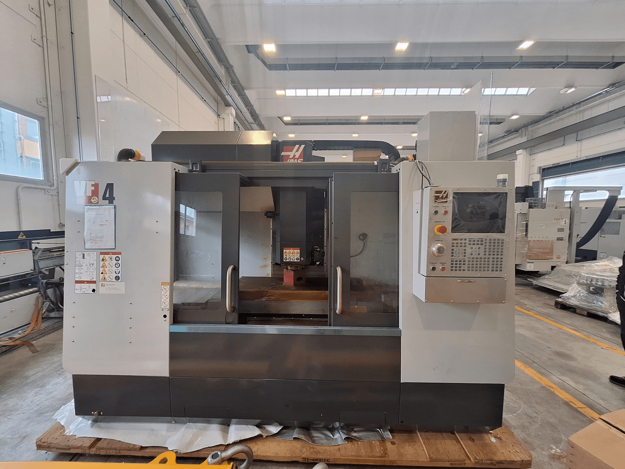 HAAS VF-4 vertikaalne töötlemiskeskus, vaade eest, millel on näha juhtpaneel, tööala ja ohutusmärgised laokeskkonna taustal.