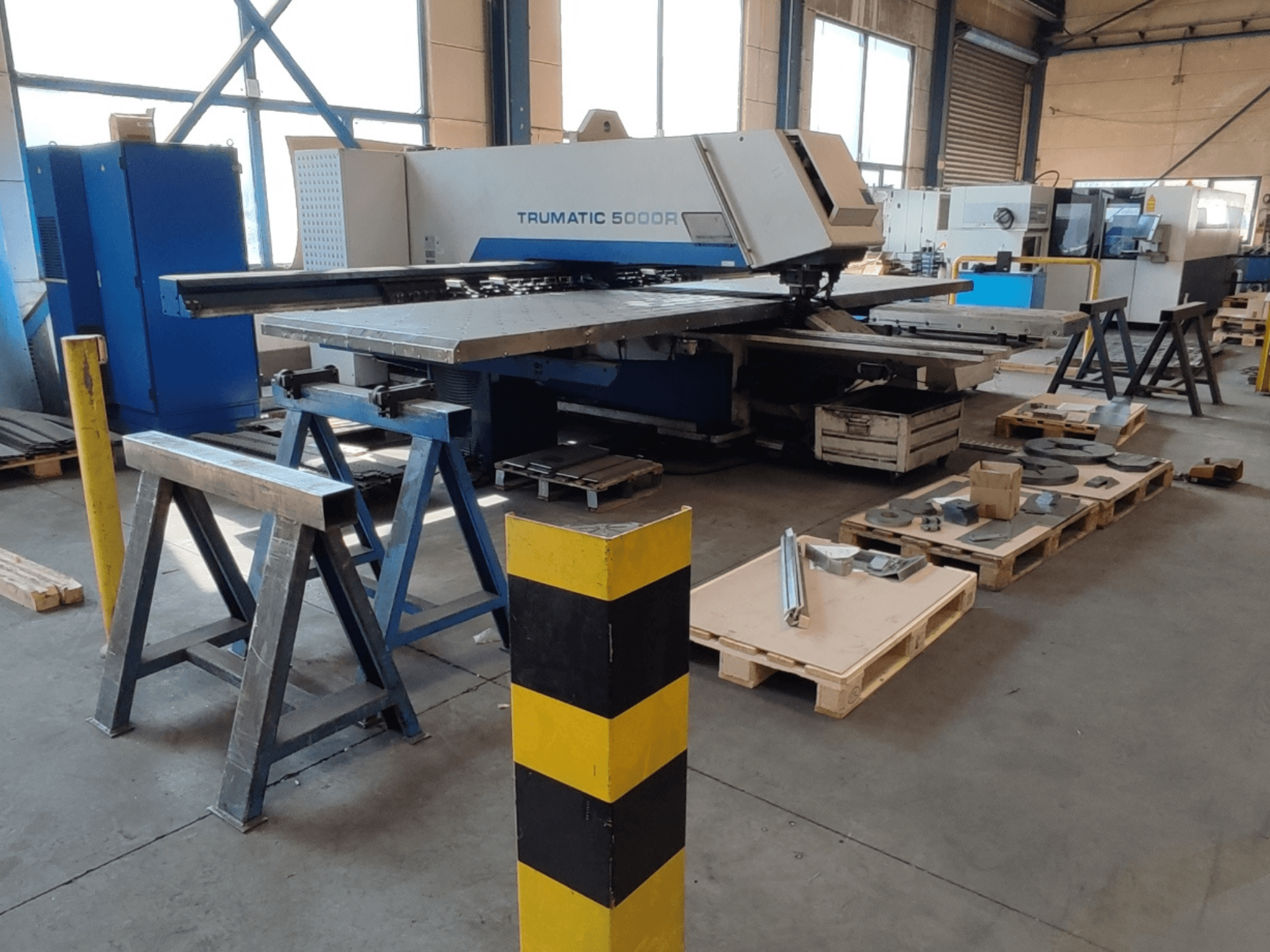 TRUMPF Trumatic 5000 R CNC-puurmasin töökodas, külgvaade, mille lähedal asuvad tööriistad ja materjalid kaubaalustel.